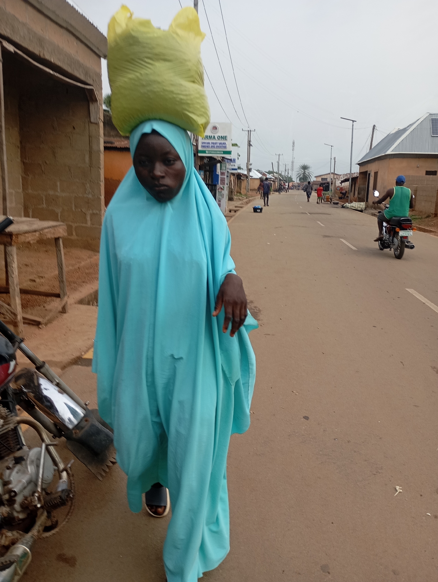 In Shendam, Nigeria, a young Black female walks along a paved road, carrying a large, bright yellow bag of goods balanced on her head. She is wearing a light blue hijab and a matching flowing blue garment that covers her body, with dark sandals visible on her feet. To her left, a motorcycle is parked on the dirt shoulder, partially obscuring her lower body. The road continues into the distance, lined with buildings and scattered with a few other individuals walking and a person on a motorcycle. The sky is overcast, suggesting a cloudy day.