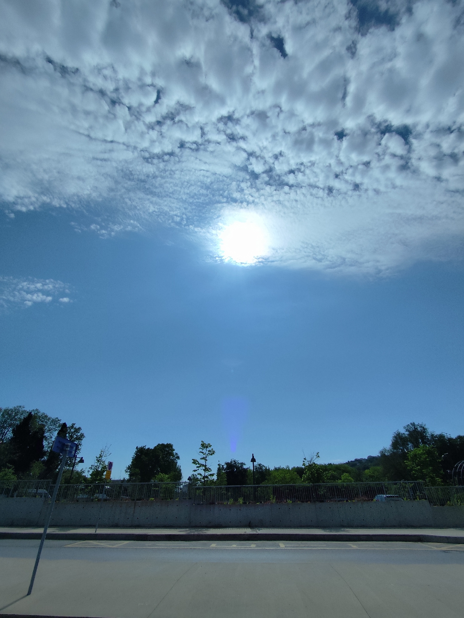 The image depicts a daytime scene in Sarıyer, Turkey, characterized by a bright, predominantly blue sky with scattered altocumulus clouds. The sun is prominently positioned in the upper center, creating a lens flare effect extending vertically downwards. Below the sky, a low concrete barrier runs horizontally across the midground, topped with a dark metal railing partially obscured by vegetation. Behind the barrier, a line of trees and shrubs is visible. In the foreground, asphalt paving is marked with faded yellow lines. A metal pole with a blue sign is situated on the left side of the road.