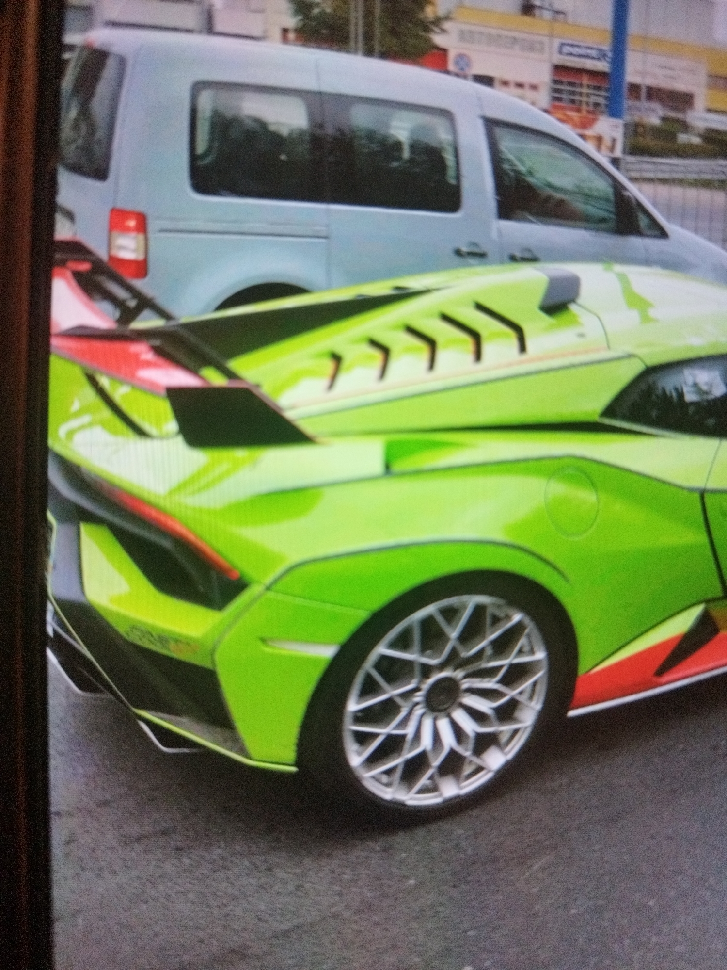 The image shows the rear portion of a lime-green Lamborghini Sián FKP 37 parked on a wet asphalt surface. The car has a distinctive rear wing, silver wheels, and black and red accents. In the background, a silver Volkswagen van is visible, and in the far distance, parts of a building are visible.
The location is identified as being within the city of Plovdiv, Bulgaria.