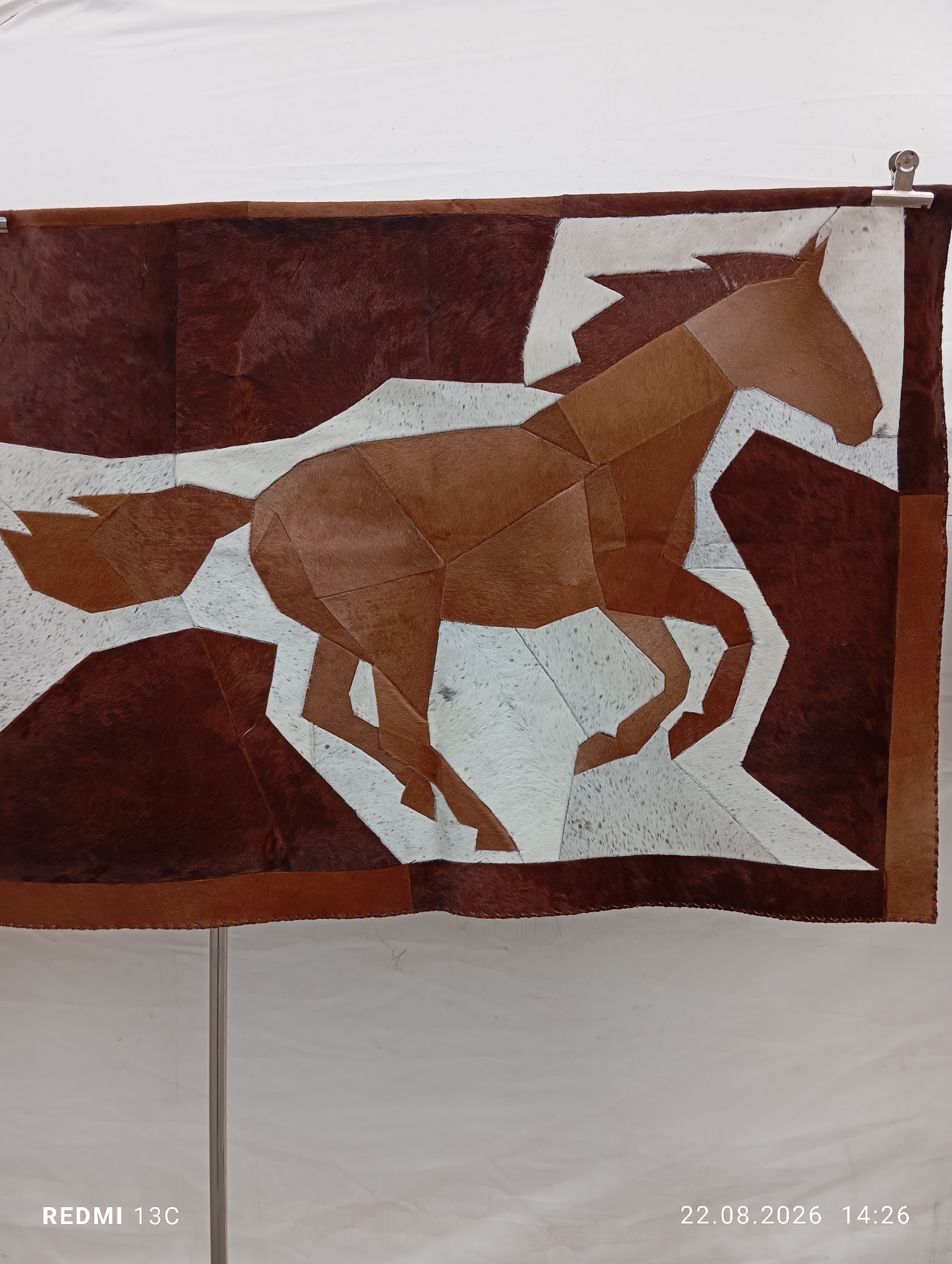 A patchwork artwork depicting a running horse is displayed against a white background. The artwork is made of leather pieces in various shades of brown and white, creating a geometric representation of the horse in motion. The horse is predominantly brown, with white areas forming parts of its body and background. The artwork is bordered by a darker brown leather strip. The artwork is attached to a rod with two clips at the top. A metal stand supports the artwork from below. The scene is set indoors with a plain white wall in the background. The bottom left corner of the image shows the text "REDMI 13C" and the bottom right corner shows "22.08.2026 14:26". There is no information about Amirsay, Uzbekistan in the image.