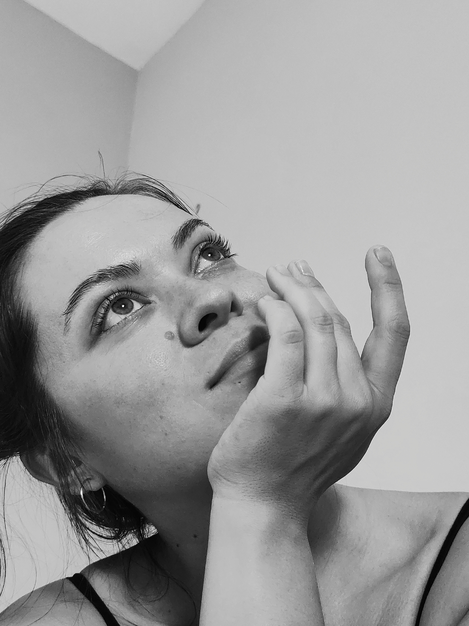 A monochrome, close-up photograph depicts a woman with her face tilted upwards, her gaze directed towards an indeterminate point above. Her hand is positioned near her face, possibly touching or resting against her cheek. The woman has dark hair, pulled back, and is wearing a tank top. An earring is visible on her left ear. The backdrop appears to be a plain wall. The photograph provides no contextual cues to ascertain the location in the city of Unknown, Unknown.