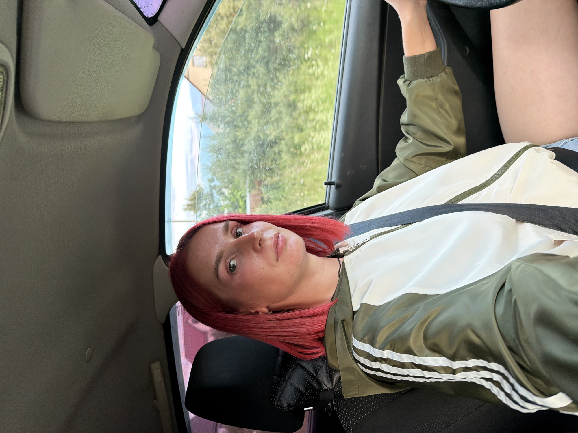 A young woman with bright pink hair is driving a car. She is wearing a green and white jacket with white stripes on the sleeves. The car's interior is visible, including the dashboard, seat, and window. Outside the window, there is greenery and a house, suggesting a rural or suburban setting. The image appears to have been taken during the daytime. The woman is looking towards the camera. Based on the license plate visible in the original image, which reads "У 264 РУ 174", the car is registered in the Chelyabinsk Oblast, Russia, which is not in the city of Chaikovsky, Russia.