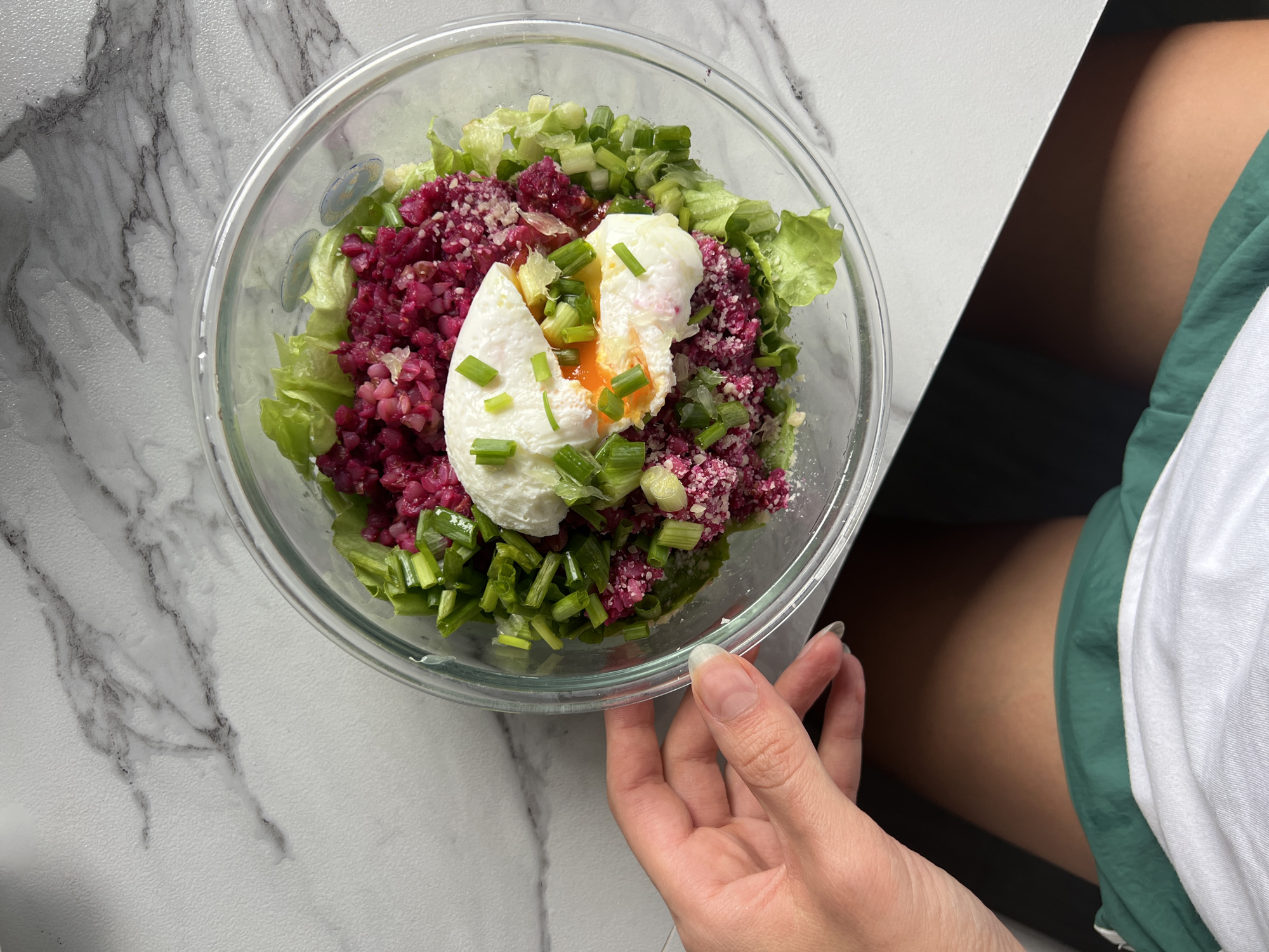 Here is a top-down shot of a glass bowl containing a salad, likely prepared at home, atop a white marble countertop. The salad consists of a bed of light green lettuce, topped with a mixture of finely chopped, dark red vegetable, possibly beets or a red cabbage. A poached egg, with its yolk visible and slightly runny, sits centrally on top of the salad. Sliced green onions are sprinkled across the salad, and a dusting of a light-colored, possibly grated, topping is present. A hand, presumably belonging to someone seated or standing nearby, is holding the bowl at the edge of the frame. The edge of a green and white garment, possibly a shirt, is visible in the upper right of the image. The location appears to be an indoor kitchen environment, most likely in the city of Unknown, Unknown.