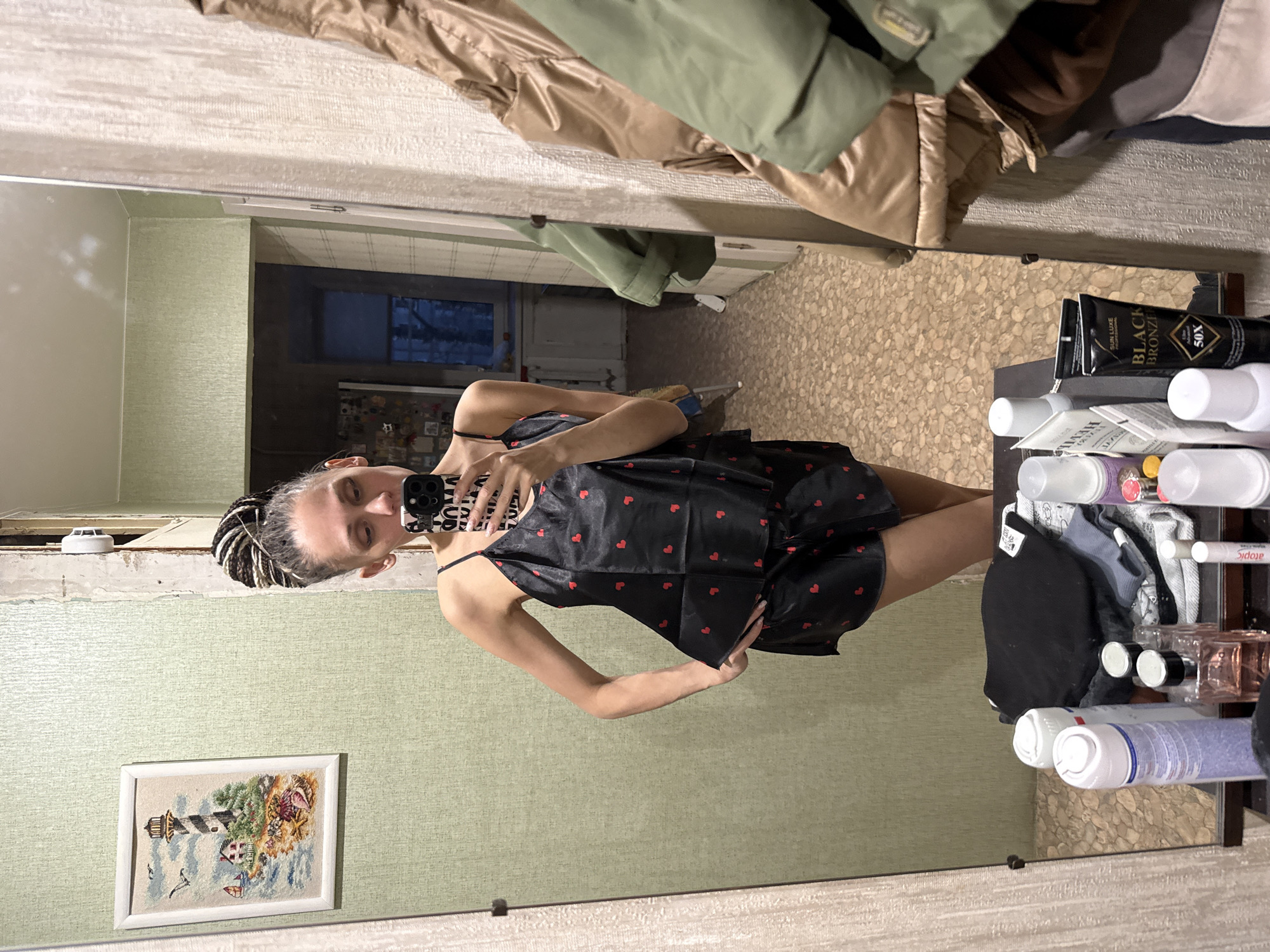 A woman stands in front of a mirror, holding a smartphone to take a selfie. She's wearing a black camisole and shorts with red hearts. Her hair is styled in black and white braids. The mirror reflects the surroundings, including a portion of a room with light green walls, a painting of a lighthouse, and a view into another room through a doorway. The mirrored reflection also shows a shelf filled with toiletries and various items. Garments are hanging above the mirror. The location appears to be an indoor domestic setting. The city and specific region are unknown.