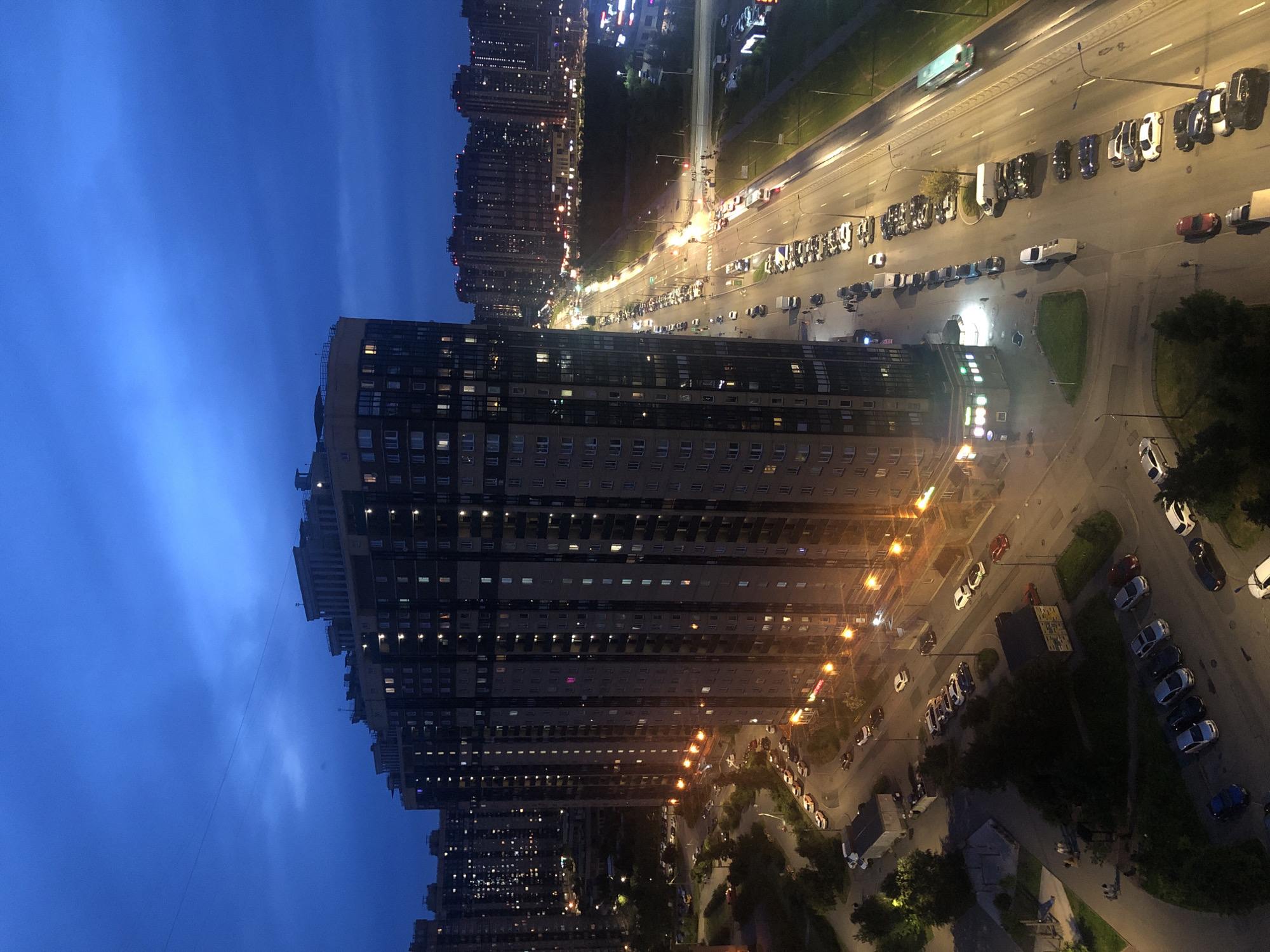 An aerial, full-frame, vertical, medium shot captures a twilight city scene. The sky is a deep, hazy blue with streaks of lighter blue and gray clouds. A complex of tall, modern residential buildings dominates the left side of the frame. The buildings are multi-story with numerous windows, many of which are illuminated with yellow and white lights. The architecture is characterized by sharp angles and a uniform, repetitive design.
In the foreground and extending into the midground are streets and parking areas. A long, multi-lane road runs from the bottom right to the upper right of the frame, lined with rows of parked cars and some moving vehicles. The road is illuminated by streetlights and the lights from the buildings. Street trees with dark green foliage are visible along the sides of some streets.
In the distance, more residential buildings of varying heights stretch across the horizon, with their windows creating a sparkling effect against the darkening sky. The overall impression is one of a densely populated urban environment at dusk or early night. There are no visible people in the frame.