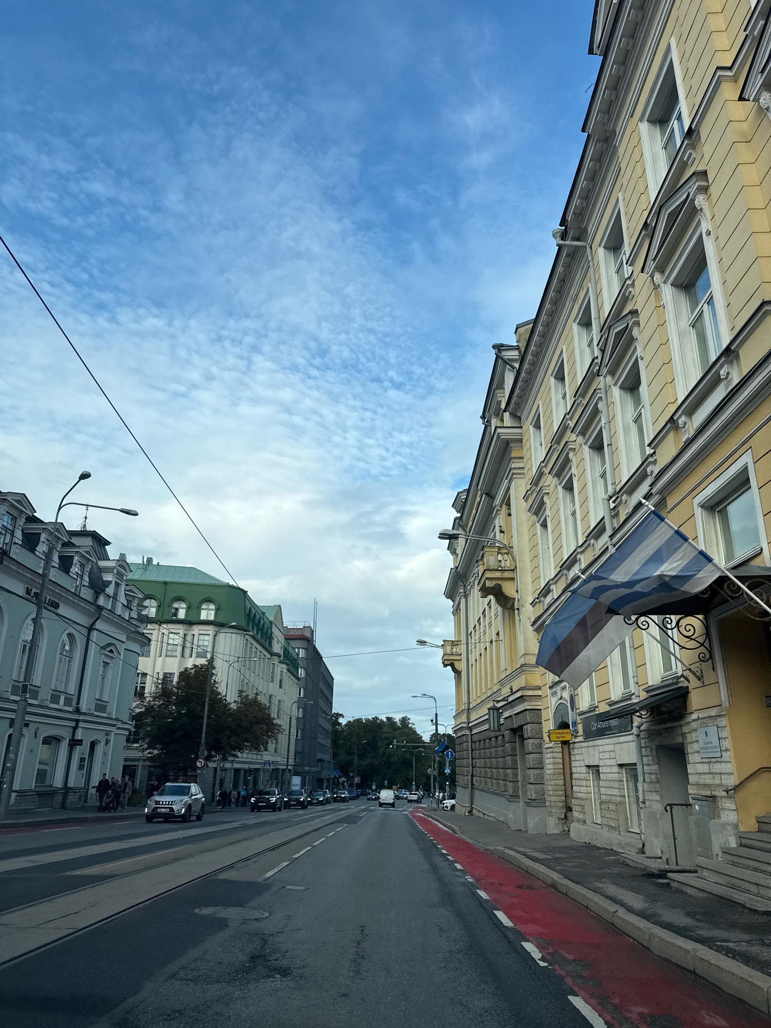 The image depicts a street scene in Tallinn, Estonia, likely within the city center. The perspective is from a moving vehicle on the road, looking forward. The street is paved with asphalt and has tram tracks running along it. On the right side of the road, there is a sidewalk with a bright red painted lane and a yellow building with multiple windows. An Estonian flag is displayed outside the building. On the left side of the street, there are several other buildings with ornate architectural details, appearing to be a mix of historical and more modern structures. The sky is partly cloudy with a pale blue hue. Several cars are visible on the road, and some pedestrians are seen on the sidewalks. Streetlights are also present. The overall atmosphere suggests a typical day in an urban environment.