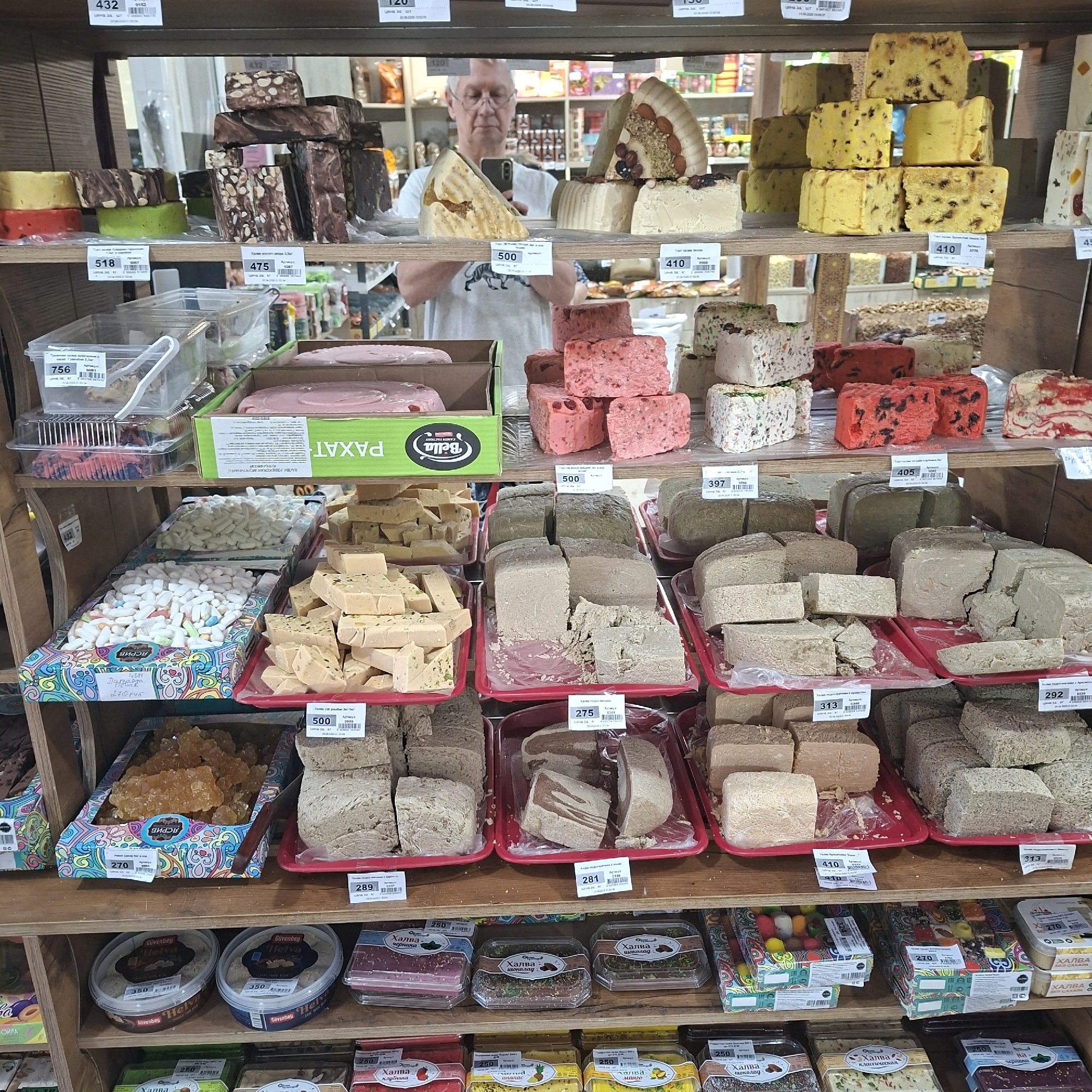 The image shows a shelf in a store, likely a confectionery or grocery store, displaying various sweets and halva. The shelves are filled with different types of halva, some in block form, others cut into pieces, and some packaged in boxes and containers. There are also other sweet items such as nougat and possibly candied fruits. The overall setting appears to be a well-stocked retail environment. A man is visible in the background, reflected in a mirror, taking a photo with his phone. He is wearing glasses and a light-colored t-shirt. The lighting suggests an indoor environment with artificial light. The items are neatly arranged on wooden shelves, with price tags visible on some of the products. The colors are vibrant, with many of the sweets having bright hues.