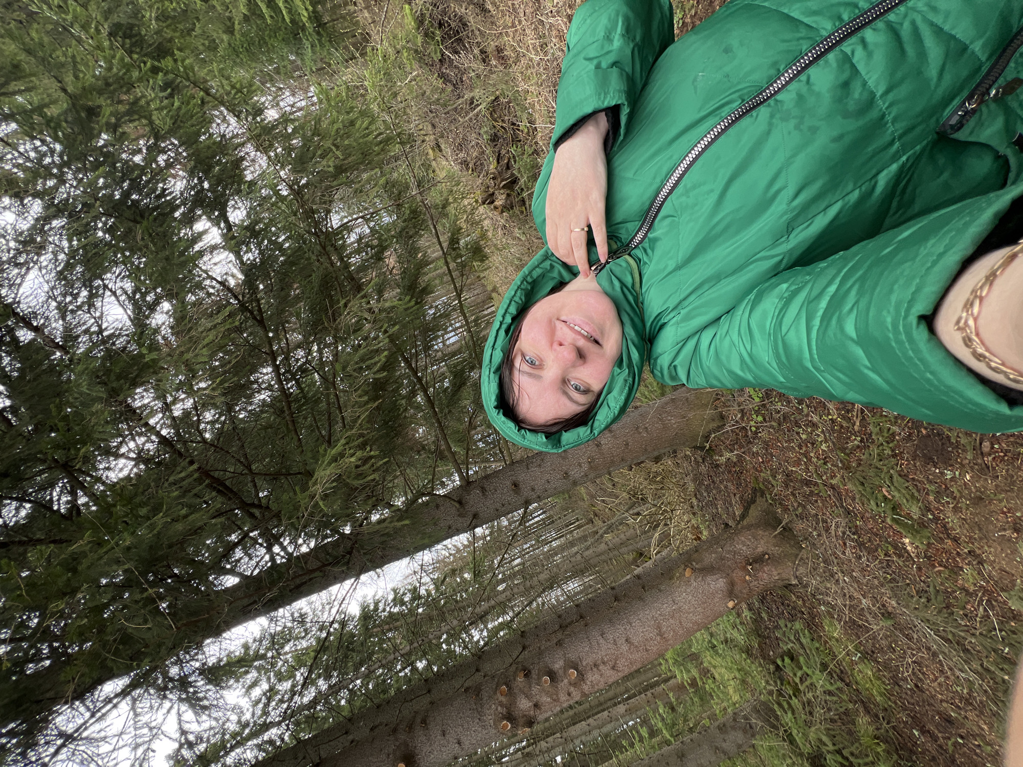 The image presents a woman in a green hooded winter coat standing in a forest. The background reveals tall, slender pine trees stretching upwards, with a view of the sky visible through the branches. The ground is covered with fallen pine needles and dry vegetation. The location is Protasy, Russia.
