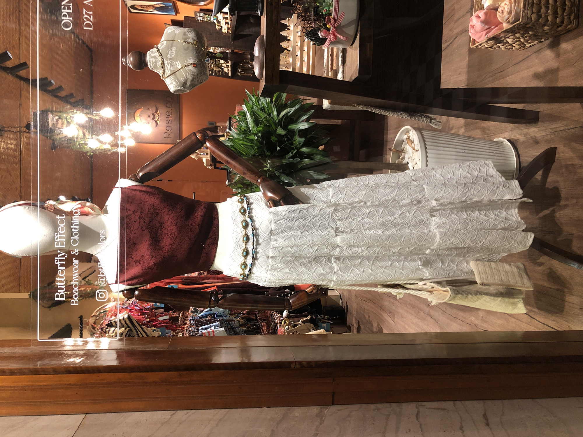 This image, taken in Da Nang, Vietnam, showcases a store window display. The primary subject is a mannequin dressed in a burgundy top and a white lace skirt, accessorized with a beaded necklace. Behind the mannequin, the store interior is visible, with shelving displaying various items, including jewelry, plants in decorative pots, and a basket filled with folded towels. A sign on the glass reads "Butterfly Effect Beachwear & Clothing" with an Instagram handle "@buterfly_effect_colors". The lighting suggests it's daytime, with sunlight reflecting on the glass. The overall impression is a well-arranged retail display designed to attract customers.