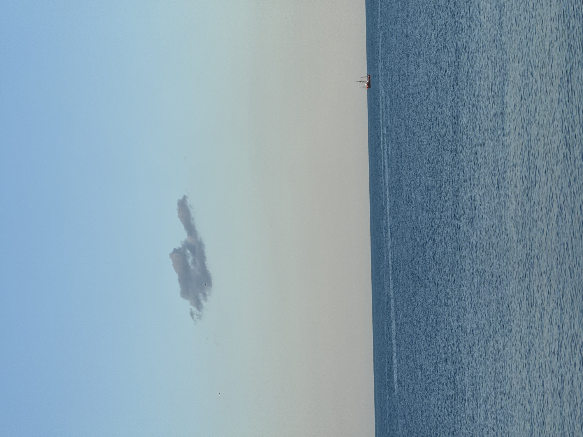 The image depicts a seascape at sea. The sky is light blue with a small cloud, the sea is blue and there is a distant ship. I do not have information to confirm the location of the image.