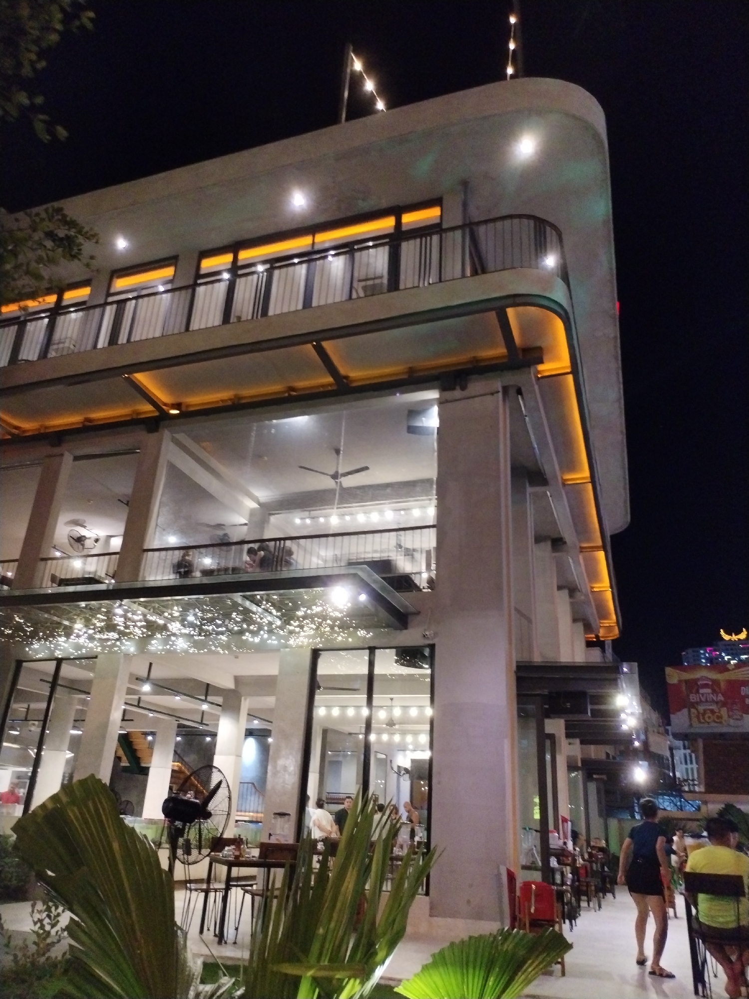 The image shows a multi-story building, likely a restaurant, in Nha Trang Ward, Vietnam, at night. The building has a modern architectural style with large windows and concrete pillars. Decorative string lights are strung along the facade, and the building is illuminated with orange and white lighting.
People are seated at tables inside the building and along the sidewalk. A large fan is visible inside. Palm-like plants are in the foreground.
In the background, other buildings and signs are visible, including one that reads "BIVINA". People are walking along the sidewalk.