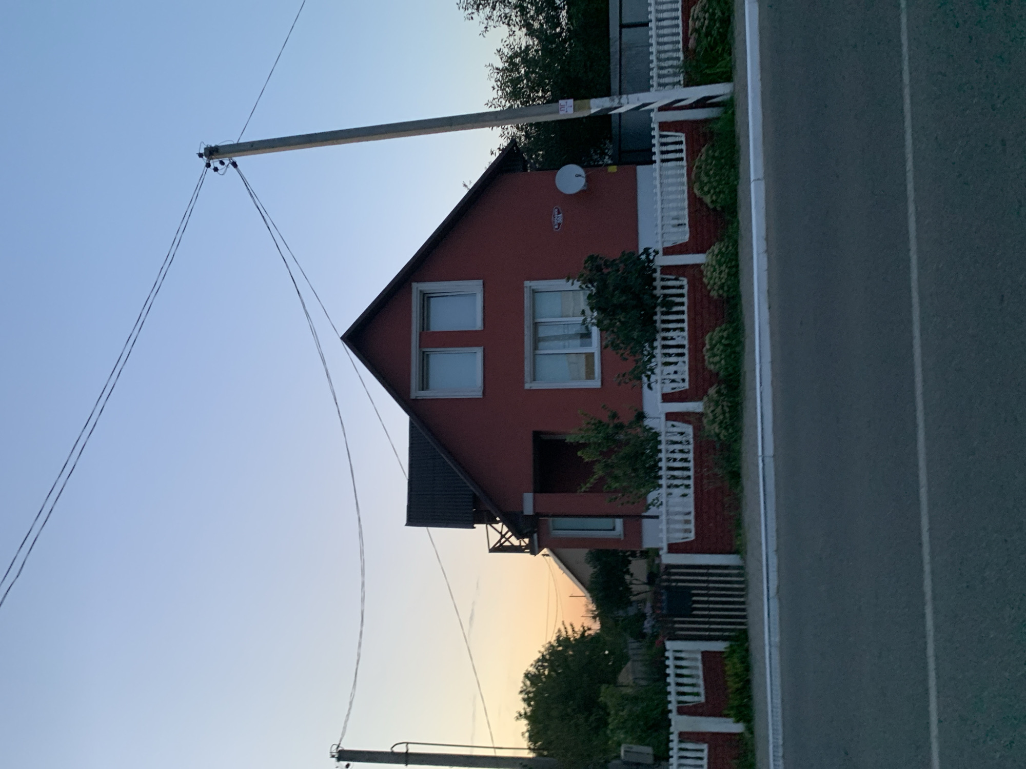 The image depicts a residential scene in Kapyĺ, Belarus, captured during the twilight hours. A two-story, red-painted house with a dark, gabled roof is the central focus, situated behind a white picket fence with a red brick base. The house features several windows with white frames, and a small satellite dish is mounted on the right side of its exterior wall. A balcony with wooden railings is visible on the second floor, on the left side of the house.
In front of the house, a paved road runs horizontally across the bottom of the frame, with a white line marking the lane. A sidewalk or curb separates the road from the property. To the left of the house, a tall utility pole with wires strung across it stands prominently. Another utility pole is visible on the right side of the image.
The environment is characterized by a clear sky transitioning from a pale blue overhead to a warm orange glow near the horizon, indicating either sunrise or sunset. Green trees and bushes surround the house, some partially obscuring the property and fence. A metal gate is positioned within the fence. The overall impression is of a quiet residential street bathed in soft, natural light. There are no people or animals visible in the frame.