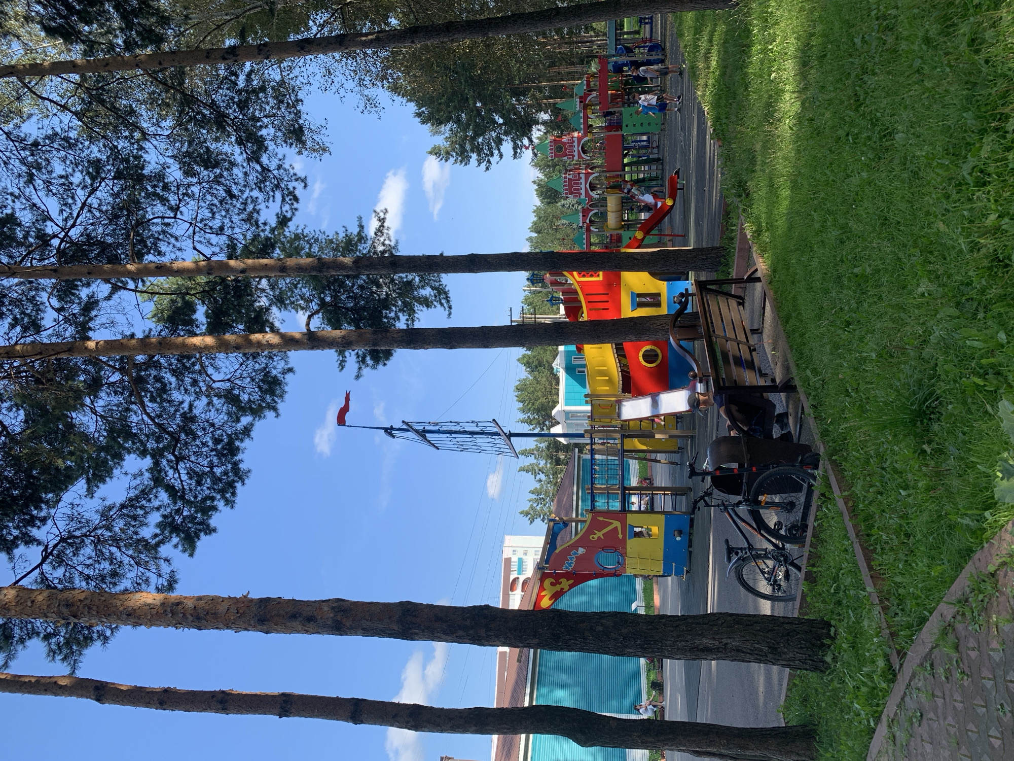 The image depicts an outdoor children's playground, likely located in Prokopyevsk, Russia, given the context. The playground features a large, colorful structure designed to resemble a pirate ship and a castle, with various slides, climbing nets, and turrets. The pirate ship section is predominantly red, yellow, and blue, with an anchor symbol. The castle section has green roofs and red accents, with a clock visible on one of the towers. Several children are playing on the equipment, including on slides and near the castle structures. Two bicycles are parked in the foreground, next to a wooden bench. A person is sitting on the bench, facing away from the camera, near a trash receptacle. The playground is surrounded by tall pine trees, casting shadows on the grass and play area. In the background, there are buildings with turquoise and brown facades, suggesting an urban or suburban setting. The sky is blue with scattered white clouds. The overall scene conveys a bright, sunny day at a recreational facility.