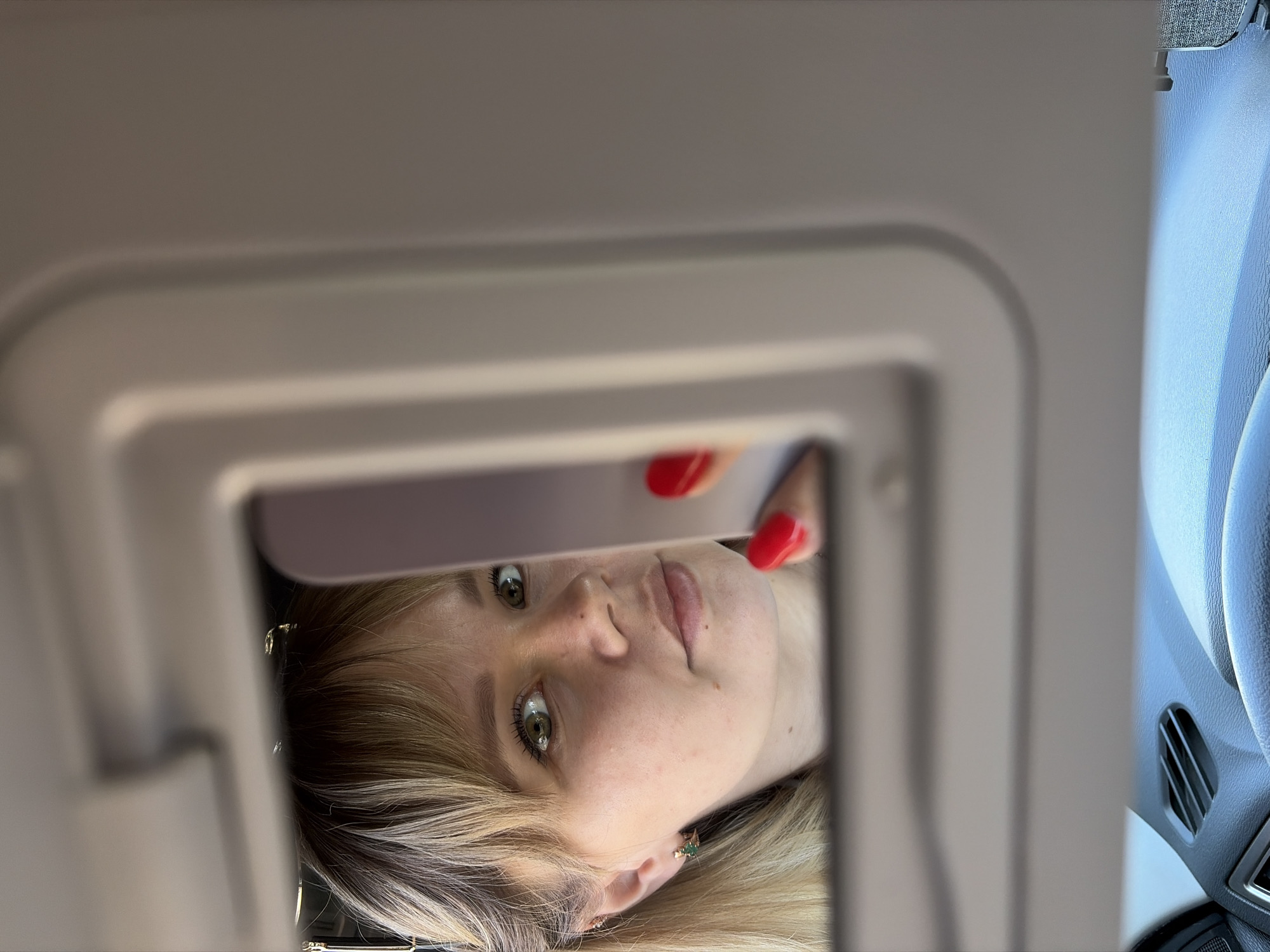 The image shows a woman's face reflected in a mirror. The mirror is part of a car's sun visor. Her hair is blonde with darker roots and she has green eyes. Her nails are painted red. To the right of the mirror is part of the car's dashboard and steering wheel. There is no information in the photo to identify the location as Chaikovsky, Russia.