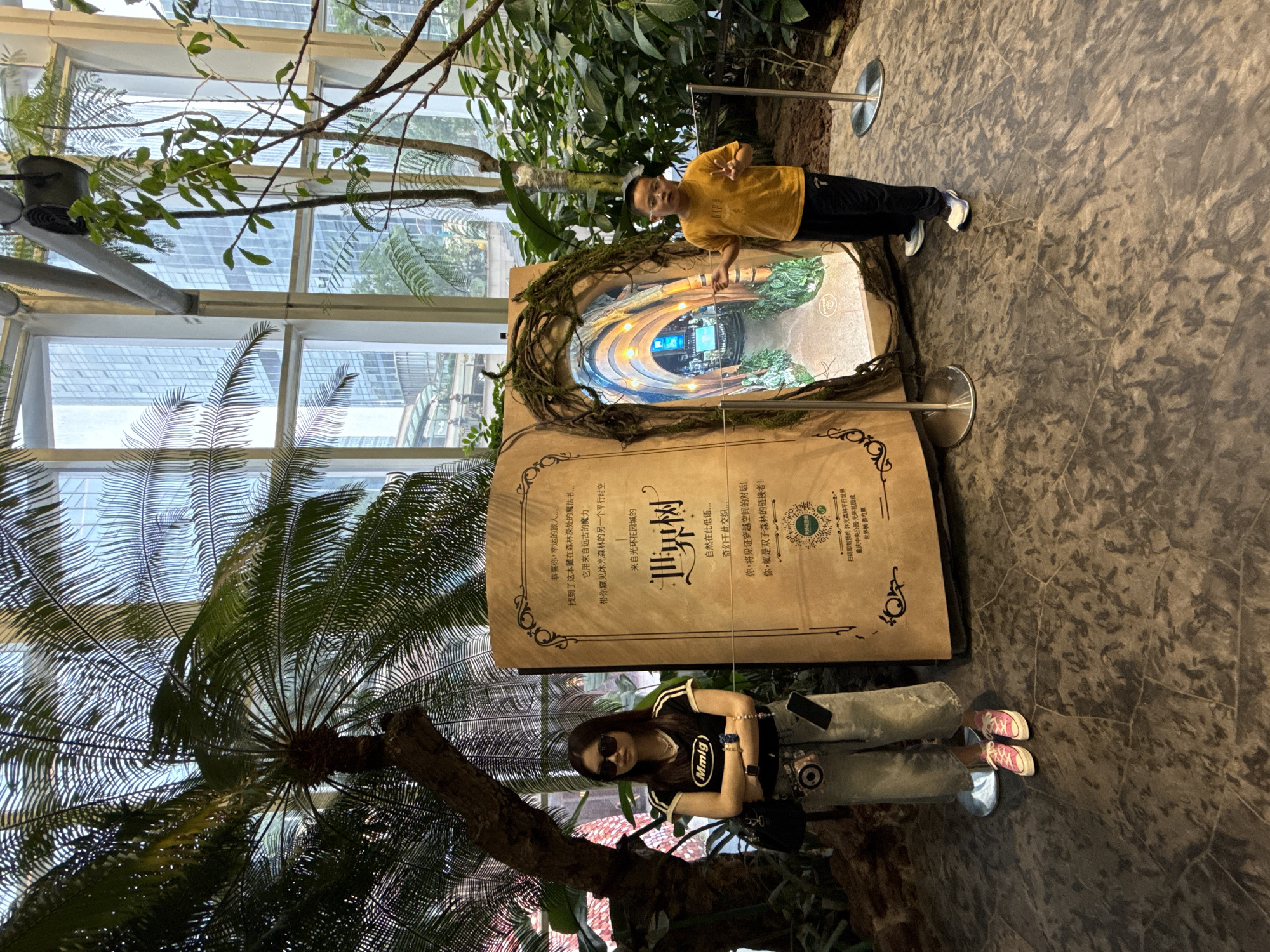 Two children stand in front of a large, open book installation in a conservatory with large windows overlooking a cityscape. On the left, a young woman wearing sunglasses, a black t-shirt with white stripes on the sleeves and a logo, light-wash distressed jeans, and pink sneakers stands with her arms crossed. A black handbag is slung over her shoulder, and a vintage camera is attached to her belt. To the right, a young boy in a yellow t-shirt and black pants, making a peace sign with his fingers, stands with his right hand extended. The book is designed to look like a portal into a fantasy world, with vines and moss adorning its opening. The conservatory features lush greenery and tall palm trees, with a modern skyscraper visible through the glass windows. A sign at the bottom right of the book display reads "扫码预约·探秘森林平行世界·重庆中央公园·时光花园·世界树·奇遇记".