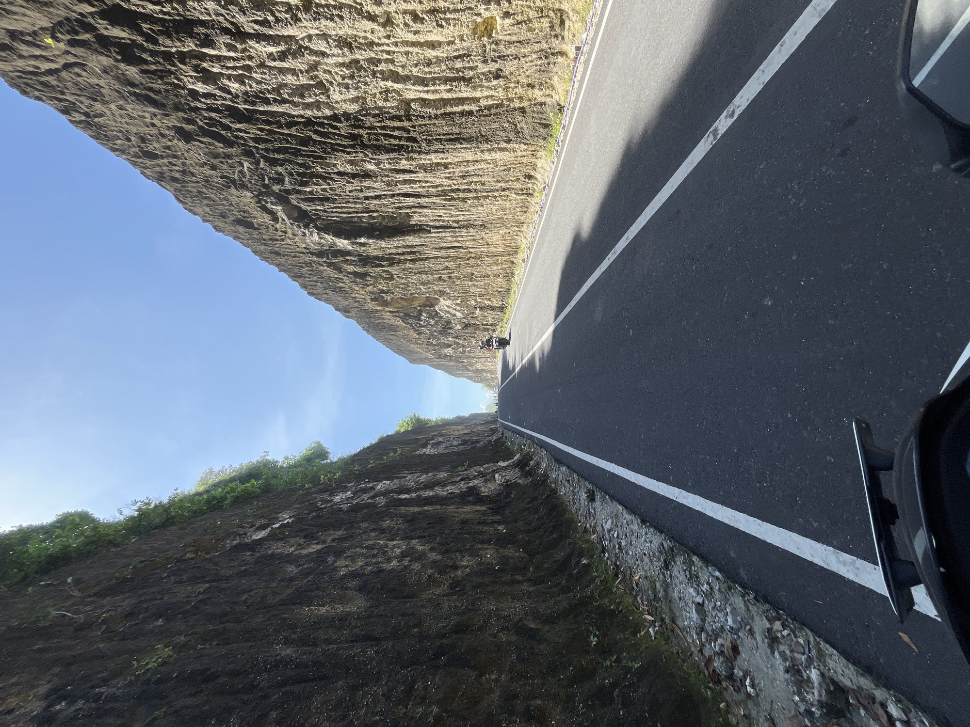 A road runs between two cliffs in Kuta Selatan, Indonesia. The road is asphalt with white lines marking lanes. A vehicle's side mirror is visible in the lower right corner. A motorcycle is seen in the distance on the road. The cliffs are composed of layered rock and vegetation. The sky is visible above the cliffs.