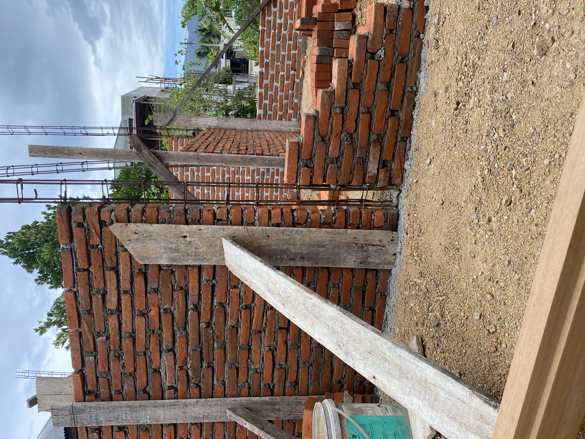 This image depicts an outdoor construction site in Banda Aceh, Indonesia. The primary subject is the partially constructed brick wall of a building. The wall is made of red bricks laid with grey mortar. Reinforcement bars protrude from the top of some sections of the wall, indicating ongoing construction. Wooden planks are used as temporary supports or scaffolding, one leaning diagonally against the brick wall. A green and white plastic bucket, likely containing building materials, is visible to the left. The ground is covered with gravel and dirt, typical of an unfinished construction area. In the background, more brick walls and some vegetation are visible, along with other buildings and a mountainous landscape under a cloudy sky. The scene suggests a construction project in progress in a tropical or subtropical environment.