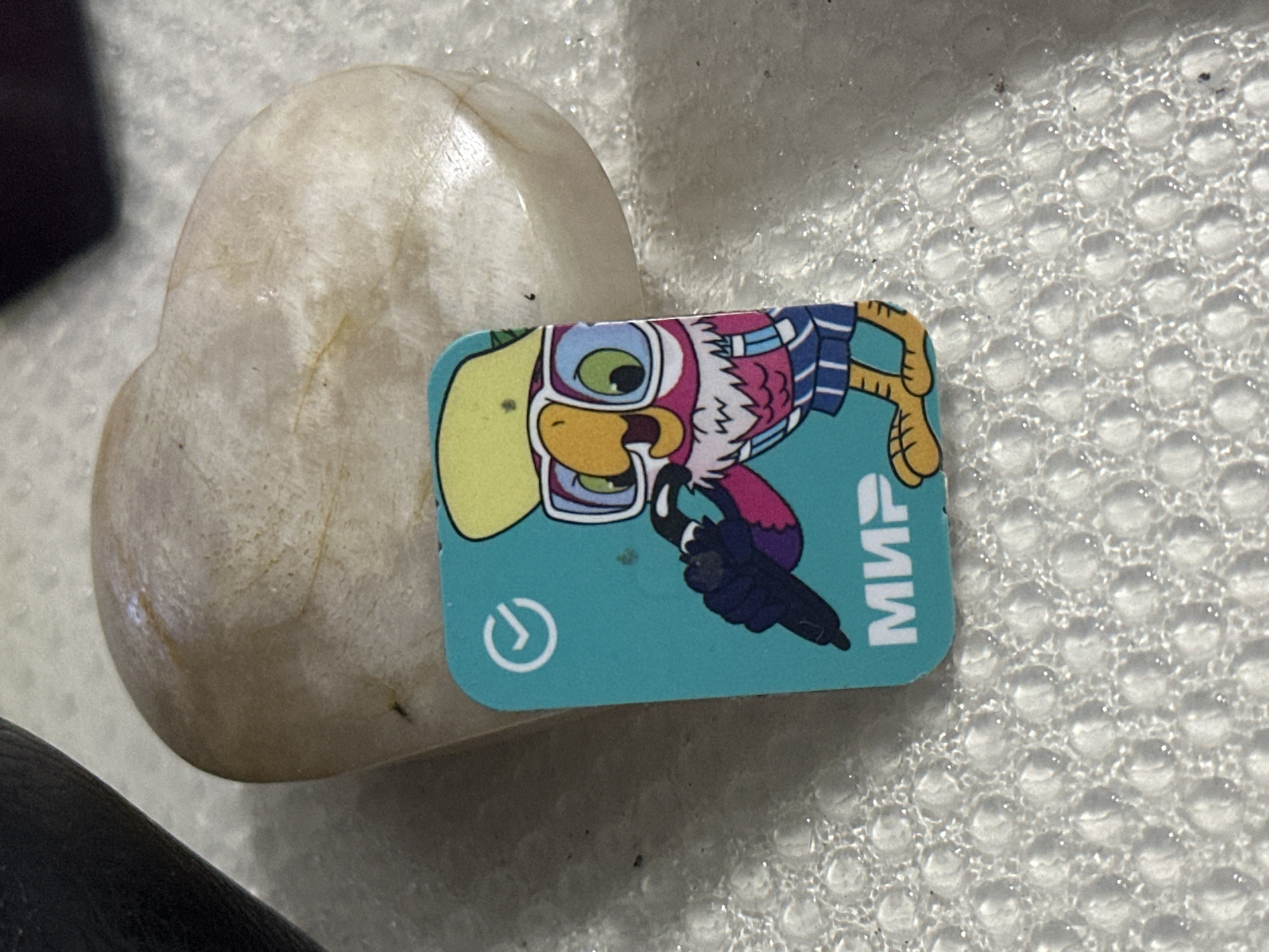 A close-up, eye-level shot shows a small, turquoise plastic card with a cartoon owl character on it, resting on a white, textured surface. The owl is wearing a yellow cap, glasses, and striped shorts, and is holding a dark object resembling a microphone. The word "МИР" (MIR) is printed in white capital letters at the bottom of the card. Behind the card, a smooth, light-colored, heart-shaped stone is partially visible. The background appears to be a textured fabric or mat. The lighting is bright, creating highlights on the stone and the textured surface. The image is devoid of any identifiable landmarks or contextual clues that would allow for its location to be determined, including within the city of Nevinnomyssk, Russia.