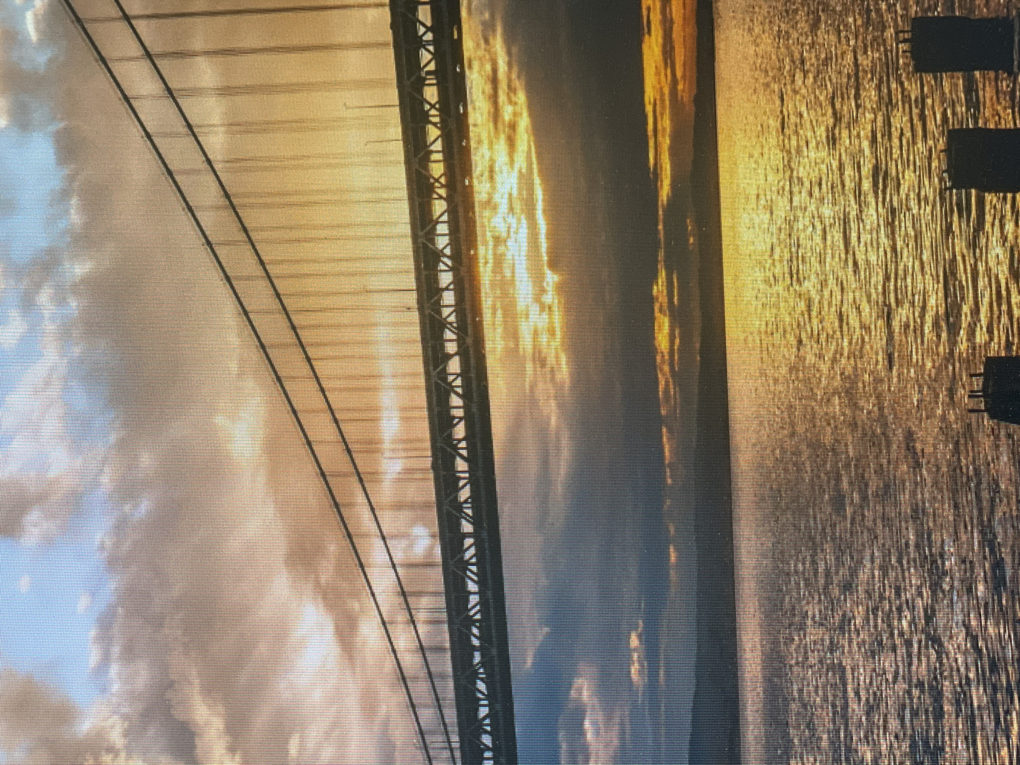 A photograph depicts a section of a bridge, likely a suspension bridge, at sunset. The bridge's suspension cables and a portion of the support structure are visible on the left side of the frame, ascending diagonally. The sky behind the bridge is filled with clouds, reflecting hues of orange and yellow from the setting sun. To the right of the bridge, a body of water, possibly a river or the sea, mirrors the golden light of the sunset. The water surface has a textured appearance due to ripples or waves. The image appears to be rotated counterclockwise. There is no evidence indicating the location in the city of Poltava, Ukraine.