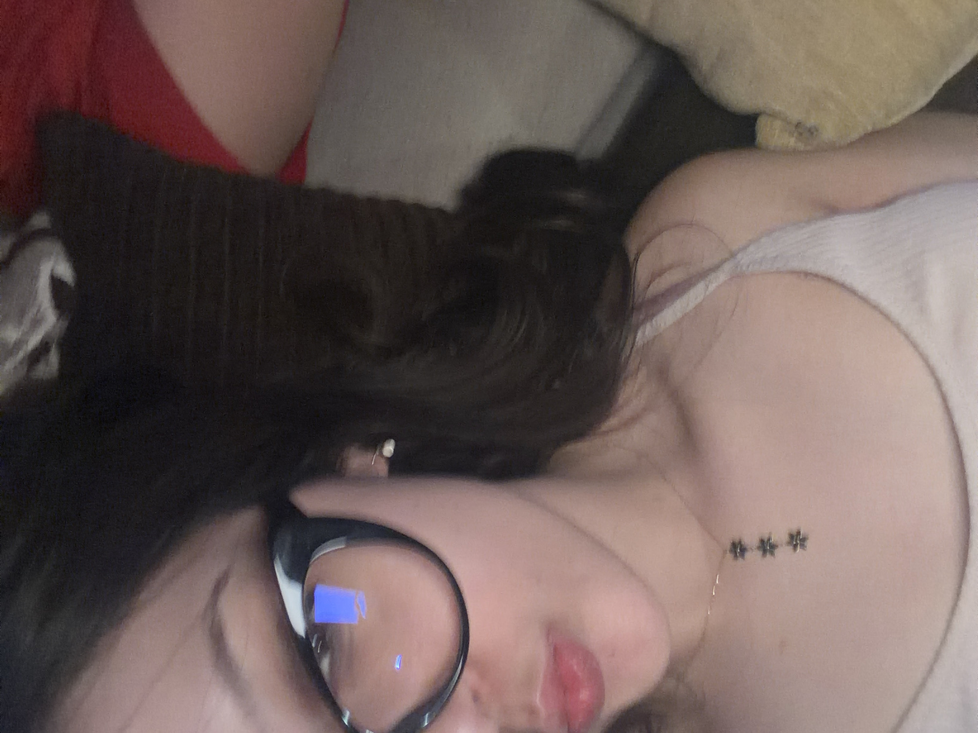 A person lies on a cushioned surface. The person is wearing glasses and a light-colored top. A dark-colored cushion or blanket is behind the person's head. The location is not specified.