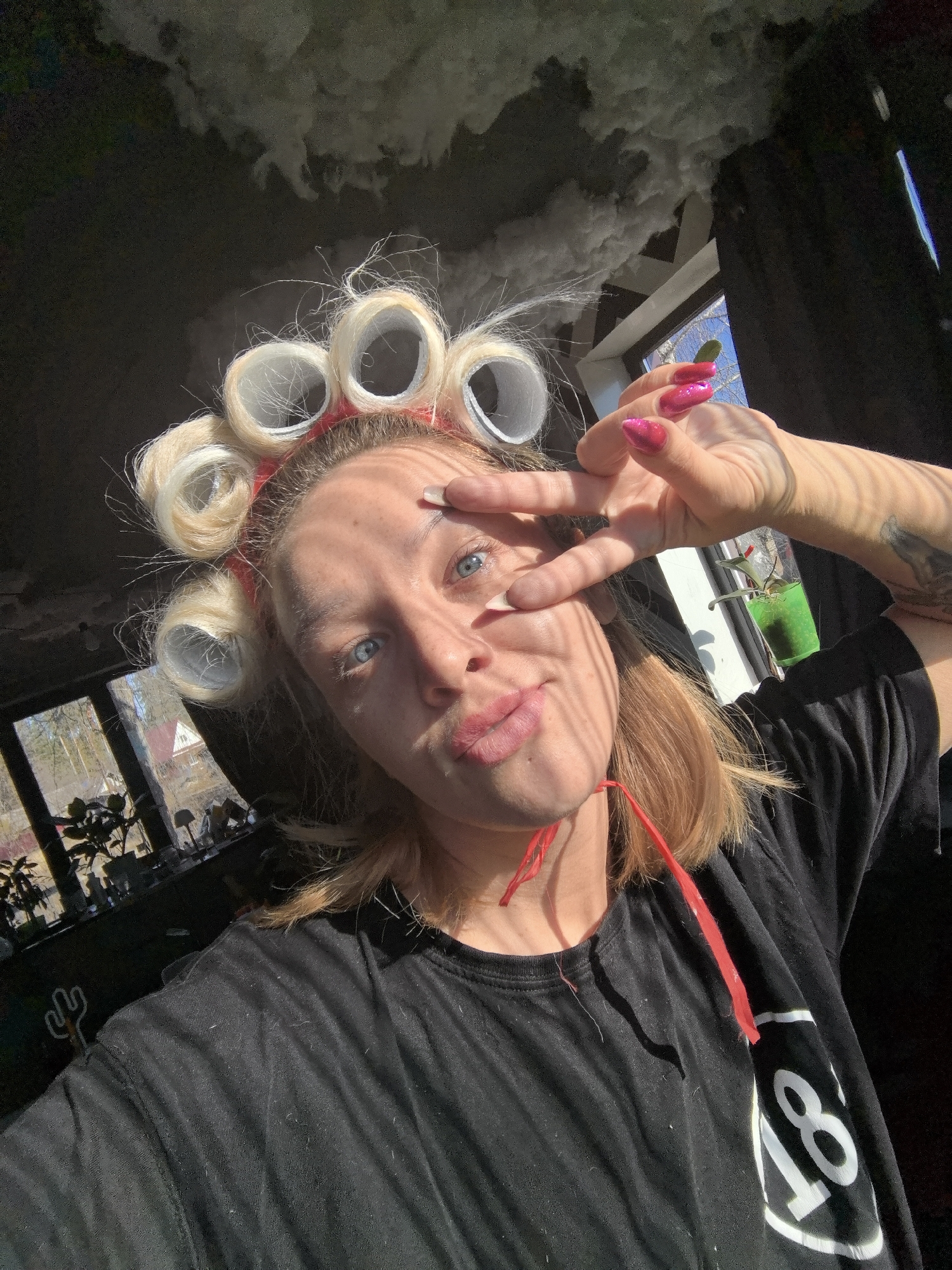 The image shows a selfie of a woman with blond hair in rollers, wearing a black t-shirt with a white number 18 on the sleeve, and giving a peace sign with her hand. She has pink glittery nail polish and a tattoo on her arm. The location appears to be indoors, with natural light coming through windows. A decorative cloud-like fixture hangs from the ceiling. Outside the windows, trees and buildings are visible, possibly indicating a residential area.