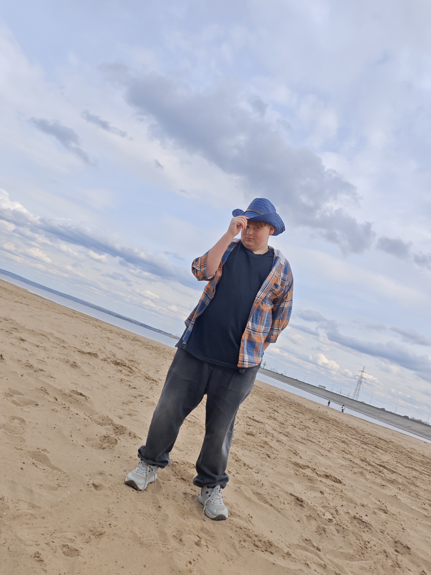 A full shot captures a young, light-skinned individual on a sandy beach beneath a cloudy sky. The person wears a blue hat, a black t-shirt, a blue and orange plaid open shirt, dark denim pants, and grey sneakers. They stand at an angle, adjusting their hat with their right hand, while their left hand is tucked behind them.
In the background, a body of water stretches towards the horizon. A long, light-colored embankment with what appears to be a road and power lines runs along the water's edge. Two small figures are visible in the distance on the embankment. The location is the city of Noviy, Russia.
