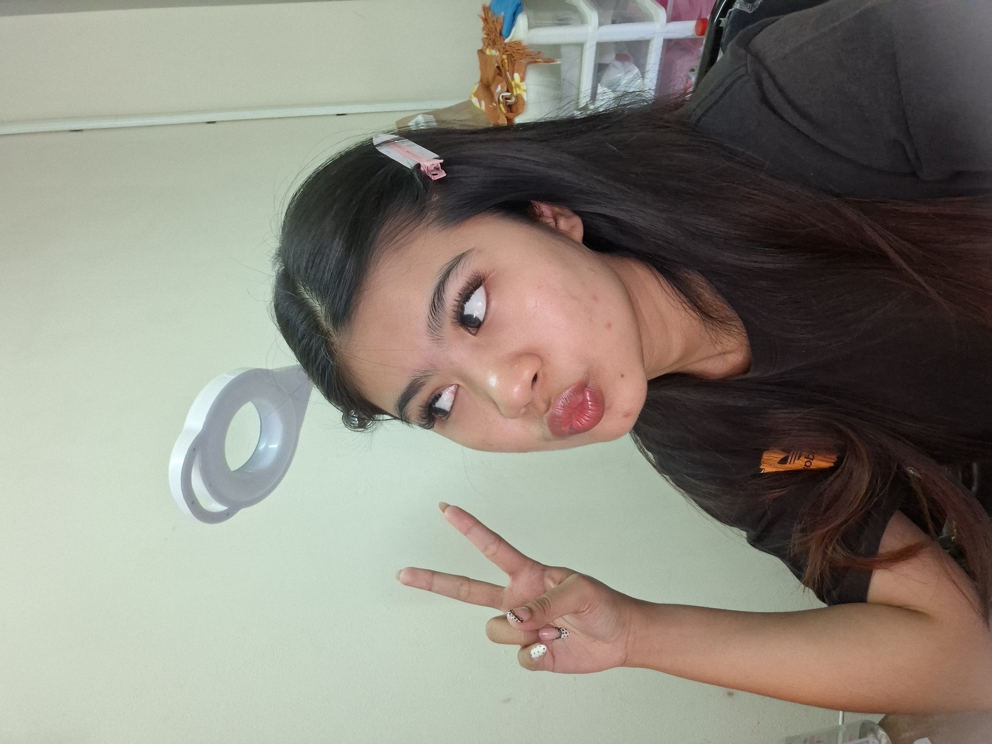 A young woman with dark hair, wearing a black t-shirt, poses for a selfie in what appears to be a home or dormitory room. She is making a peace sign with her left hand, which has decorative nail art. Her lips are pursed, and she is looking upwards and to her left. A white ring light is visible in the background, suspended from the ceiling. To the right of the woman, there are shelves with various items, suggesting a living space. The walls are a light, neutral color. The location is identified as Kuta, Indonesia.