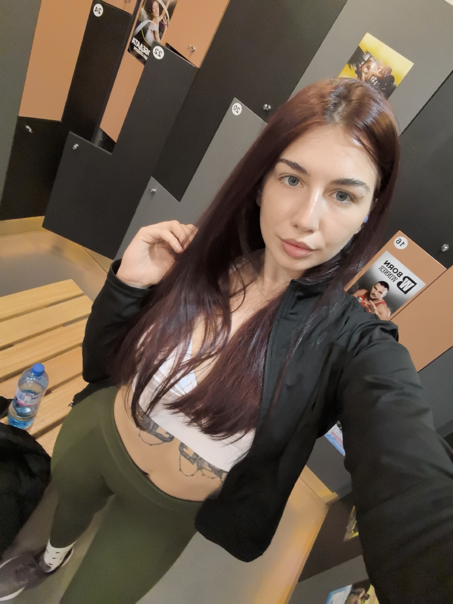 A medium shot depicts a woman with long, auburn hair in a locker room setting. The woman is fair-skinned with light eyes. She is wearing a white sports bra, olive-green leggings, and a black zip-up jacket. A tattoo is visible on her midriff. She is holding her hair with her right hand.
Behind her, the backdrop consists of lockers, some with numbered labels and promotional posters. A wooden bench is to her left with a water bottle placed on it. The image was taken in the city of Stara Zagora, Bulgaria.