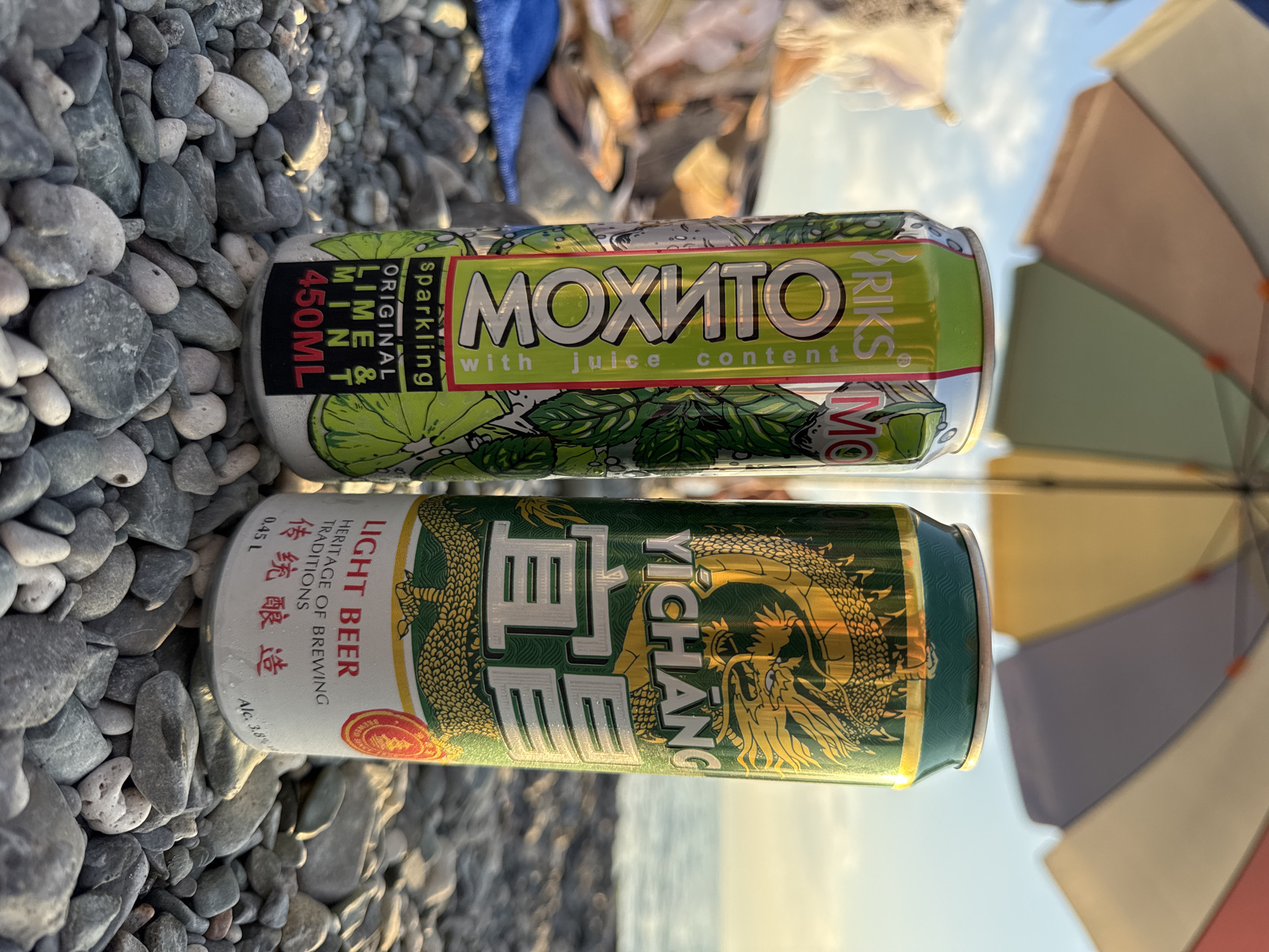 Two beverage cans, a green "MOXITO" sparkling lime and mint drink and a green "YICHANG" light beer with a dragon design, are positioned on a pebble beach in Zharbulak, Kazakhstan. The cans are partially obscured by an open beach umbrella, which casts shadows on them. The background shows a calm body of water and a cloudy sky. There are no people visible in the image.