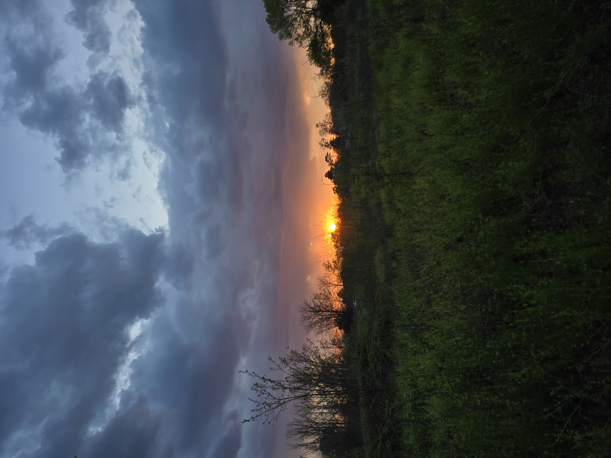 The image shows a sunset with dark clouds on the left and green vegetation on the right. The sun is positioned near the horizon, emitting a bright orange glow. There are trees silhouetted against the sunset. The location is Unknown, Unknown.