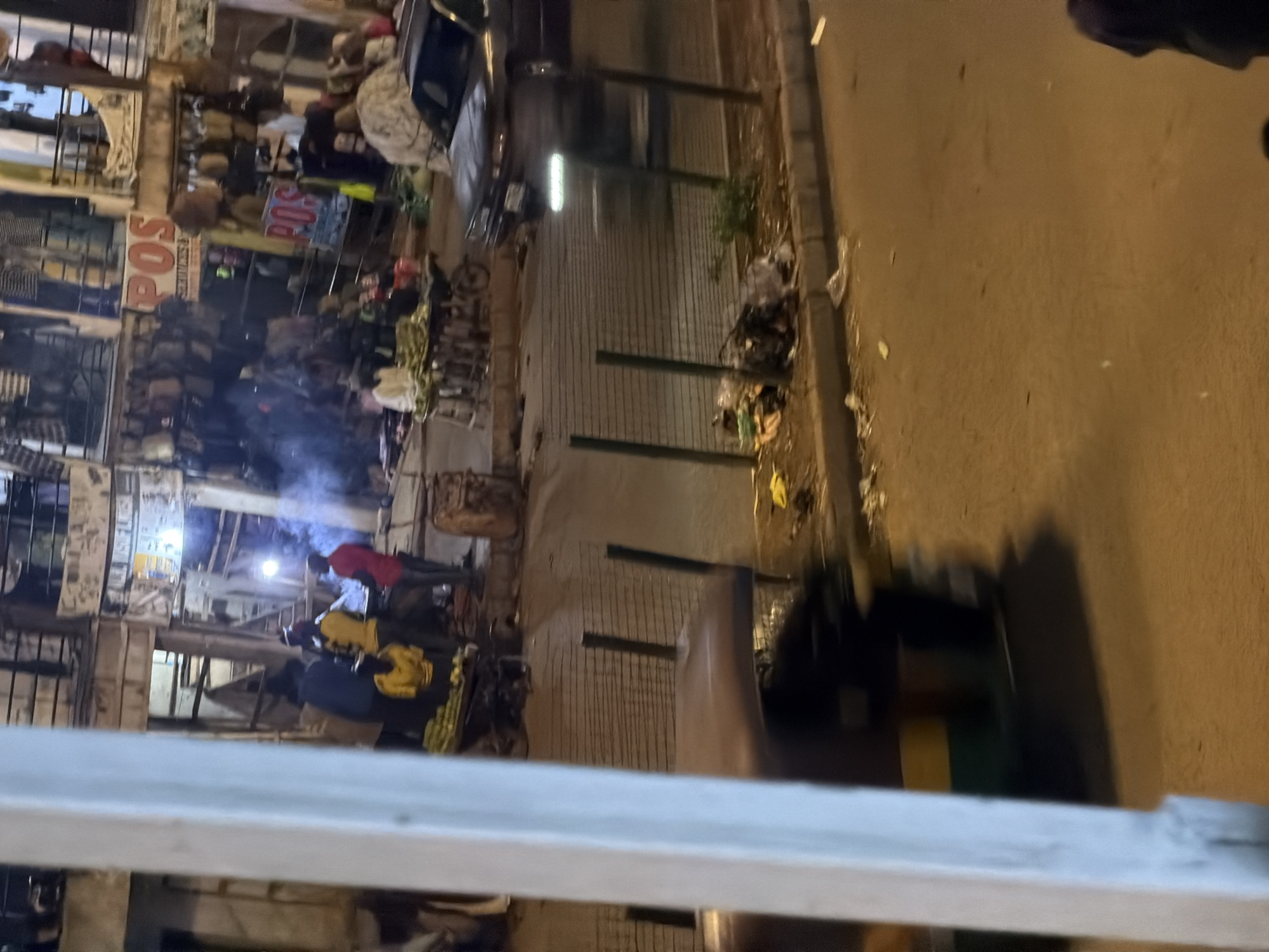 The image depicts a street scene in Bukuru, Nigeria, at night. In the foreground, a blurred auto-rickshaw with green and yellow colors is moving from left to right. A white pillar is prominent on the left side of the frame. Beyond the auto-rickshaw, a metal fence separates the street from a sidewalk area. On the sidewalk, there is a pile of trash.
Further back, on the right side of the image, a dark-colored sedan is parked. In the background, a bustling street market is visible. Stalls display various goods, including bags and clothing. Several people are present in the market area, some appearing to be vendors and customers. Two red and orange signs with the word "POS" are visible on the stalls. The market is illuminated by artificial lights, creating a lively atmosphere despite the late hour. The overall scene suggests a typical urban environment in Nigeria, with active commerce and street life.