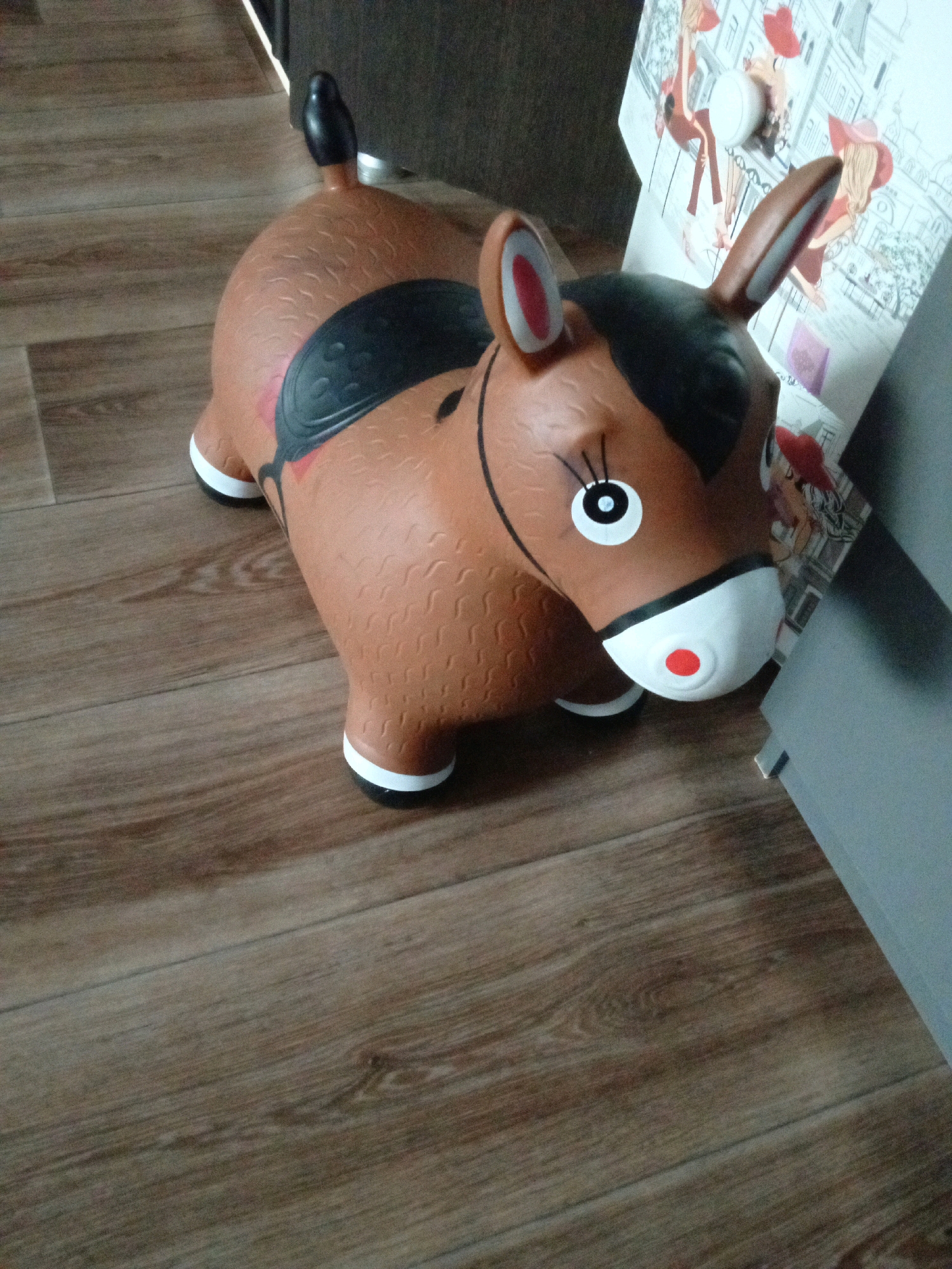 A brown, inflatable horse toy is positioned on a wooden floor. The toy has black ears, a black saddle, and a white muzzle with a red dot. A picture with cartoonish figures is visible in the background, leaning against a gray surface. The scene is indoors, likely in a residential setting. There are no people present in the image. The lighting suggests an indoor environment with artificial light. The floorboards are laid horizontally and have a wood-grain pattern.
