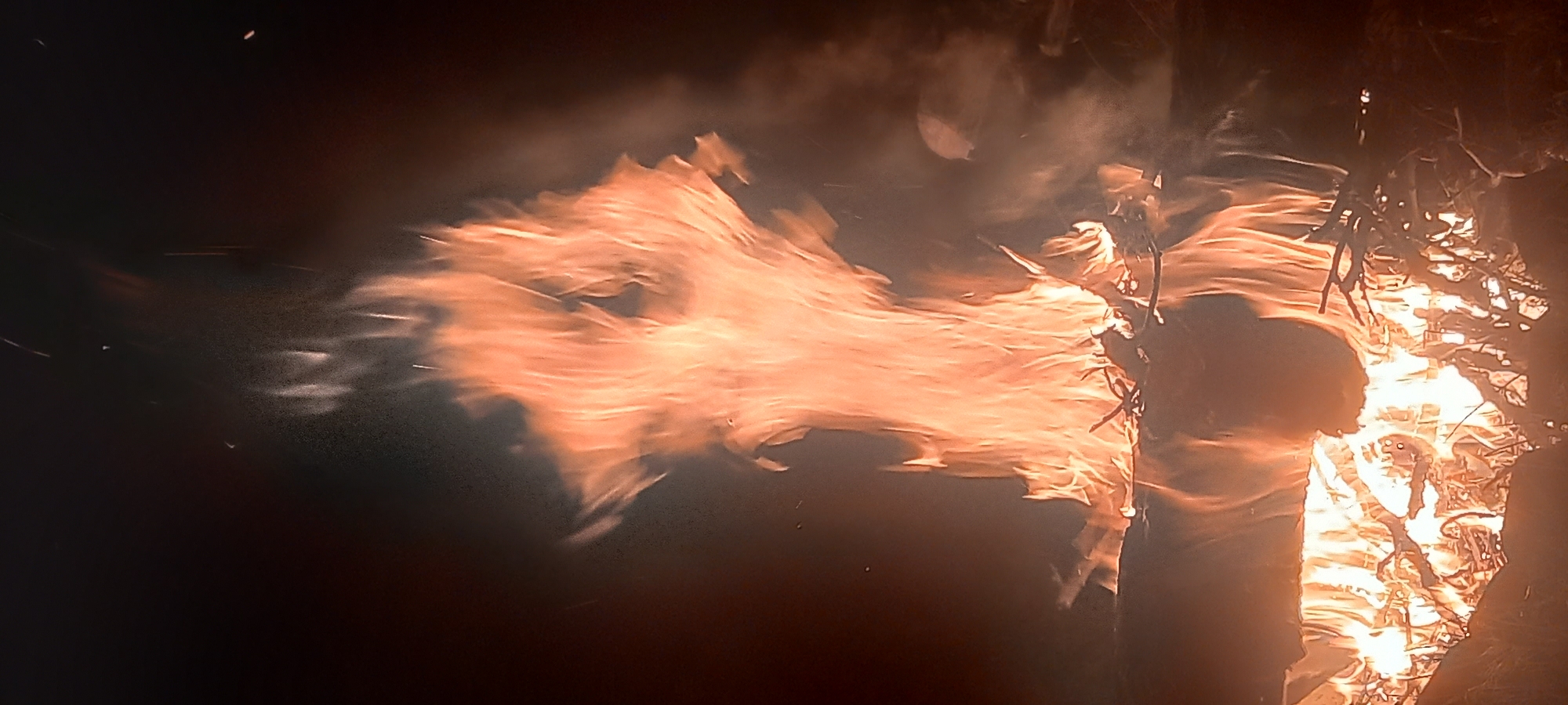 The image displays a vibrant bonfire observed at night. A large volume of orange and yellow flames dominates the central and upper portions of the frame, exhibiting an upward, turbulent flow. Significant luminescence emanates from the fire, illuminating the immediate surroundings. At the base of the flames, visible fuel consists of burning logs and numerous smaller branches. Multiple bright, upward-streaking sparks and individual glowing embers are discernible against the dark background, particularly in the upper half of the image, indicating active combustion. Some translucent smoke plumes are intermingled with the flames, particularly on the right side. The background is uniformly dark, providing no discernible environmental features or structures. The image does not contain any visual elements or metadata to identify its specific location within the city of Kryvyi Rih, Ukraine. No individuals or additional objects are visible within the frame.