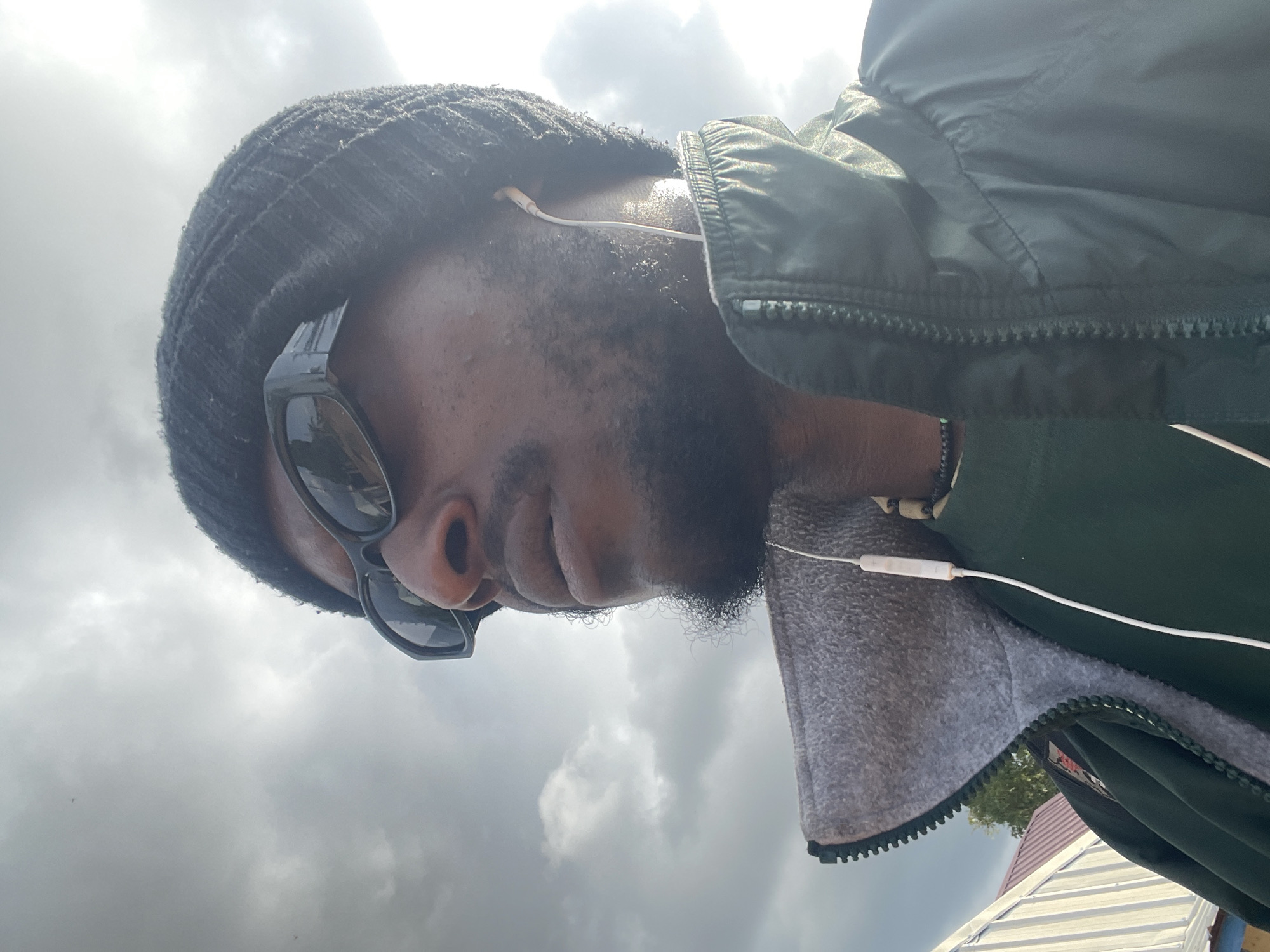 A close-up, profile shot captures a dark-skinned person wearing a black beanie, dark sunglasses, and a dark green jacket with a grey collar over a dark green shirt. White earbuds are visible, with one in the ear. The person has a beard and a necklace with dark beads and a green pendant. The background consists of a cloudy sky, with some structures and trees visible on the left. The scene appears to be outdoors.