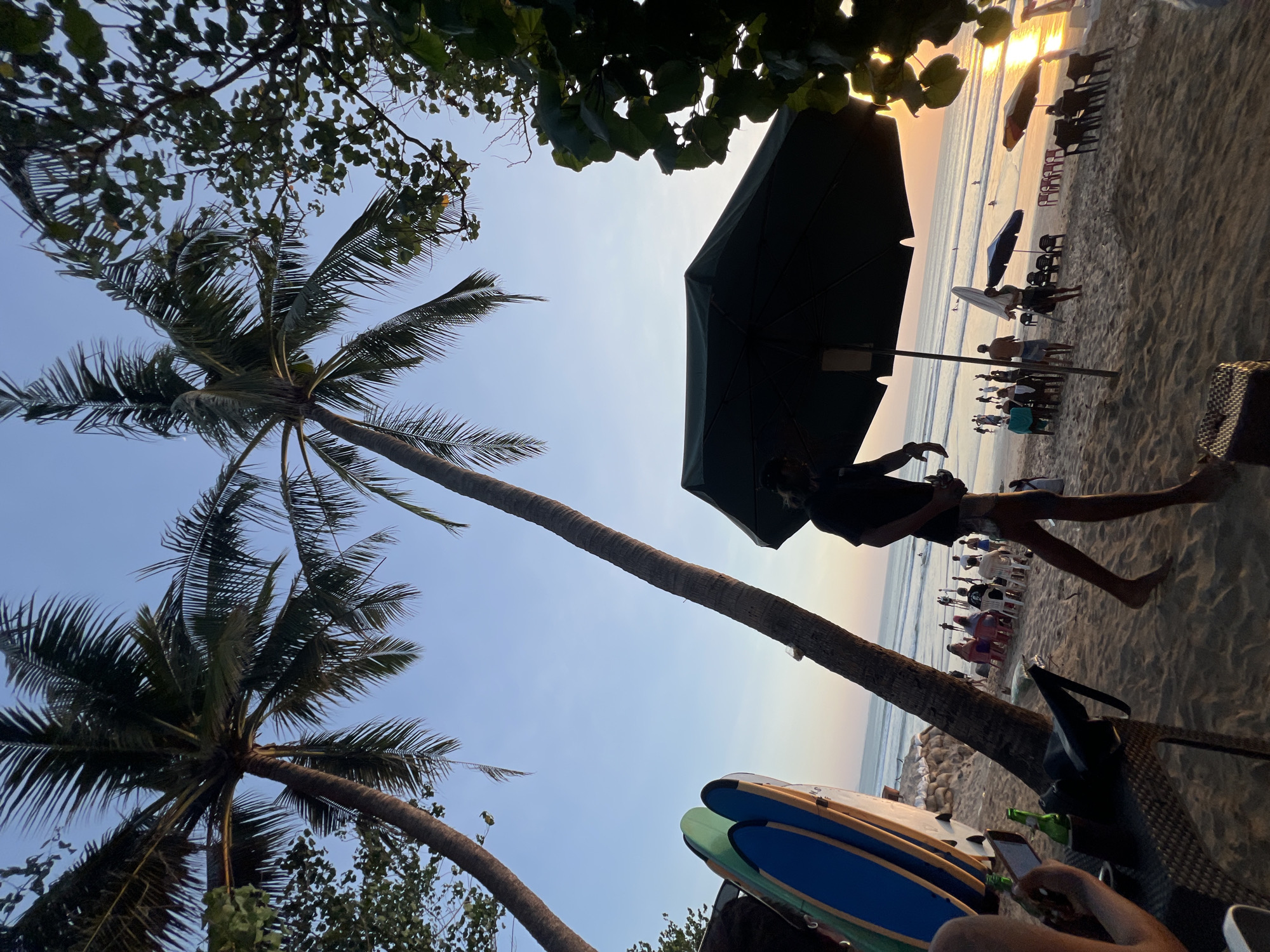 The image depicts a beach scene in Kuta, Indonesia, likely during sunset. Two prominent palm trees frame the sky, with their fronds casting shadows on the sand. In the foreground, a table holds two beer bottles and a phone, suggesting a relaxed atmosphere for patrons. To the left, two surfboards stand upright, leaning against what appears to be a sandbag wall.
Further into the scene, several people are scattered across the beach. Some are seated on chairs, possibly at beachside cafes or restaurants, while others are standing or walking. In the distance, the ocean meets the horizon, with small waves breaking and a few individuals visible in the water. Several beach umbrellas, some open and others closed, are dotted across the sand, indicating a place for beachgoers to seek shade. The overall ambiance suggests a leisurely evening at a popular tourist destination.