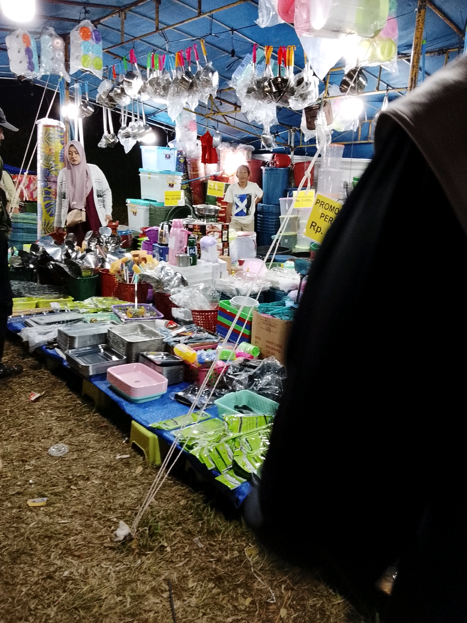 A bustling outdoor market stall is captured at night, likely in Padang Dalom, Indonesia, as evidenced by the Indonesian currency (Rp.) displayed on signage. The scene is filled with a variety of household items, predominantly kitchenware and storage containers, displayed on tables covered with blue tarps. In the foreground, a person wearing a black garment is partially visible, their back turned towards the camera. To the left, a woman wearing a light pink hijab and a grey cardigan stands near a stall, looking towards the merchandise. Behind her, a colorful cylindrical object, possibly fabric or decorative material, is on display. Hanging from the ceiling, numerous ladles and other kitchen utensils are displayed in plastic packaging. The ground is covered with dry leaves and scattered debris, indicating a somewhat informal market setting. A bright yellow sign with red lettering advertises a promotion, reading "PROMO PER Rp. 25.000". The lighting suggests artificial illumination from overhead bulbs, creating a warm and inviting atmosphere despite the darkness of the night. Various plastic containers, baskets, and bags of goods are stacked and arranged for sale.