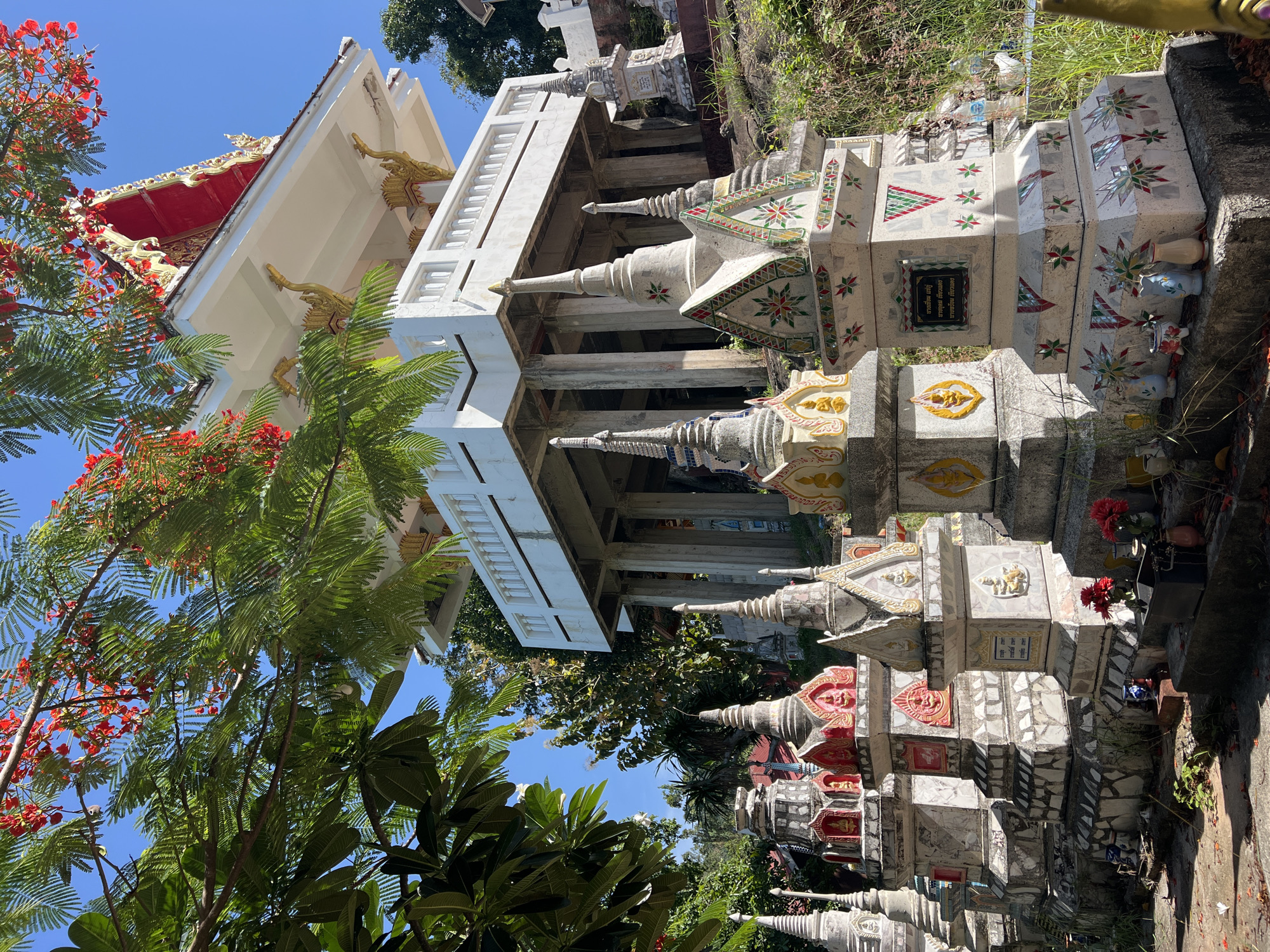 In Baan Hua Thanon, Thailand, a Buddhist temple complex is depicted under a clear blue sky. The foreground features a collection of intricately decorated stupas, each adorned with mosaic tiles in geometric patterns and Buddhist iconography. Some stupas have niches containing Buddha images. Small offerings of flowers and vases are placed at the base of the stupas. Behind these structures, a multi-tiered temple building rises, supported by numerous pillars. The temple has a white exterior with a red roof and ornate golden decorative elements. Lush green foliage and branches from a flowering tree with red blossoms frame the scene from the left and above, casting dappled shadows on the stupas. The overall environment is serene and spiritual, bathed in bright sunlight.