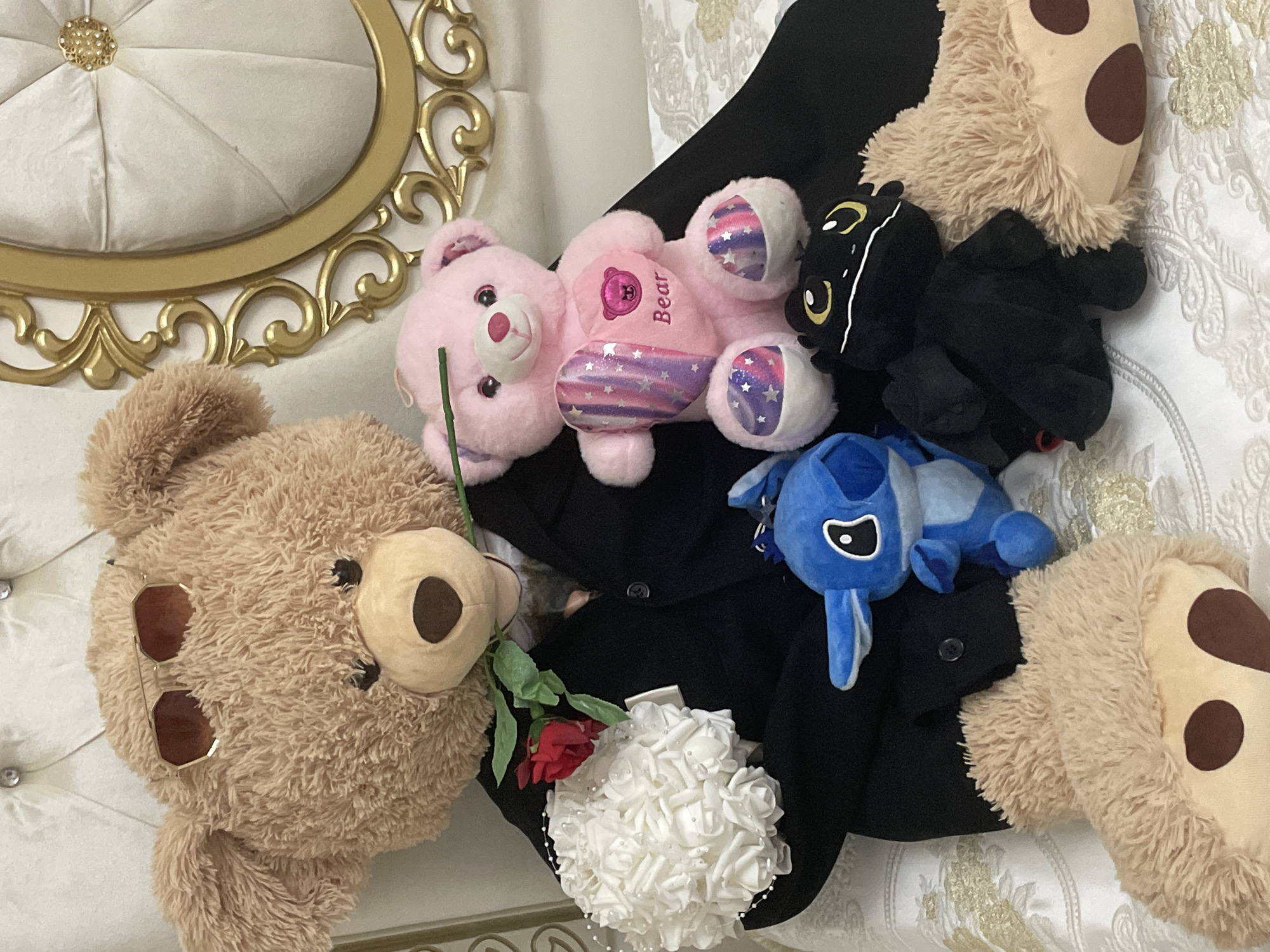 The image displays an indoor scene, likely in Ihtiman, Bulgaria, featuring an arrangement of plush toys on luxurious furniture. A prominent large, light brown teddy bear is positioned on the left, lying on its back with sunglasses resting on its head and holding a red rose with a green stem. Next to it, a pink teddy bear lies on its back, with the word "Bear" embroidered on its tummy and its paws adorned with sparkly, star-patterned fabric in pink, purple, and blue.
Among the plush toys, a black stuffed animal with yellow eyes and white accents, possibly a cat or dragon, is visible, alongside a blue plush toy resembling Disney's Stitch. Part of another large light brown teddy bear, with spotted paws, is seen on the right. These toys are primarily arranged on a spread-out piece of black fabric, possibly a jacket or suit. A small white bouquet of fabric roses, embellished with tiny beads, is placed near the red rose.
The background includes ornate furniture: a white tufted surface with a central golden button and elaborate gold scrollwork on the left, suggesting a fancy headboard or chair. On the right, a white fabric with an embossed or embroidered gold pattern is visible, likely bedding. There is no specific activity taking place beyond the arrangement itself.