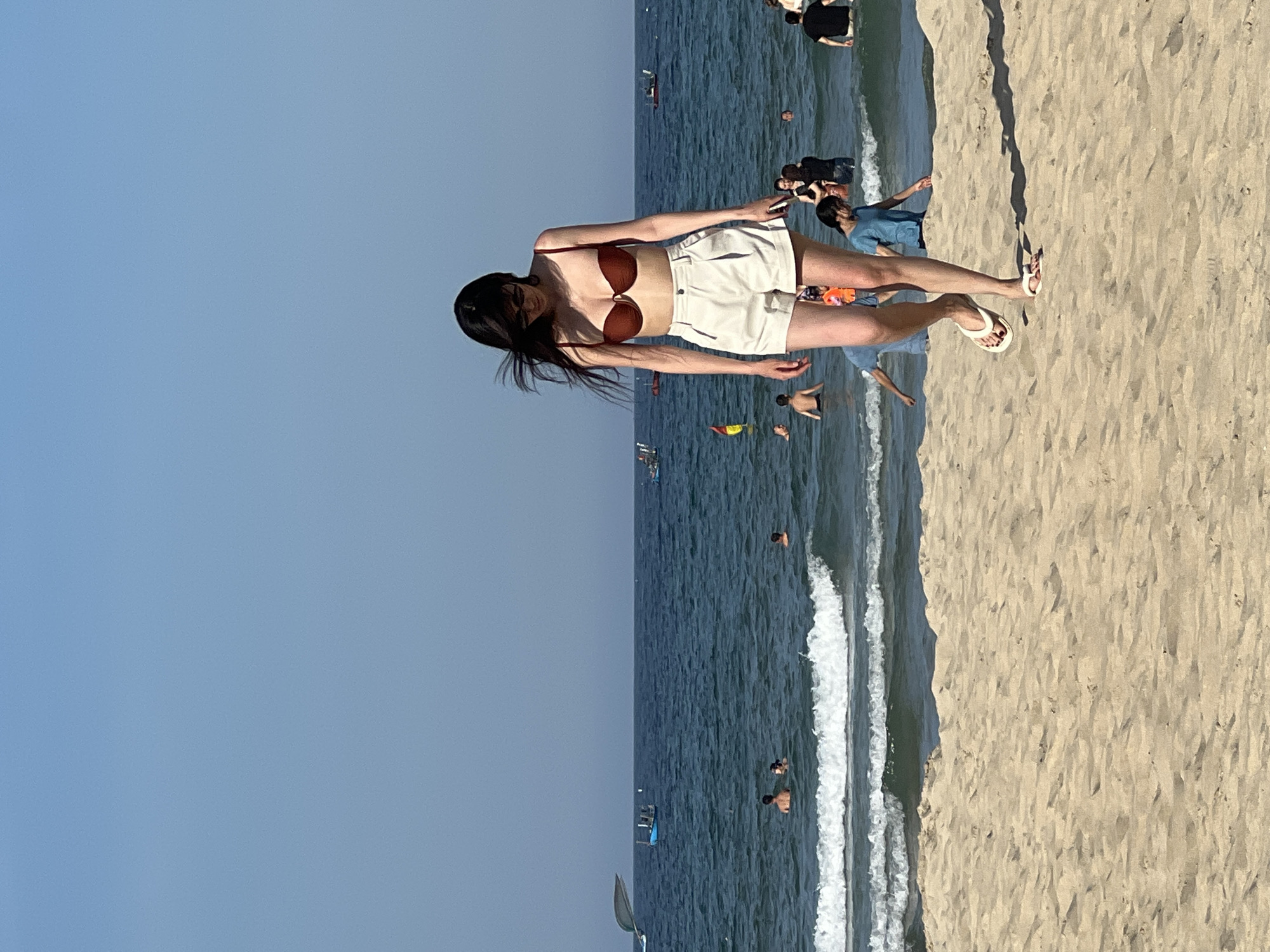 The image shows a sunny day at a beach in Da Nang, Vietnam. A young woman with dark hair, wearing a reddish-brown bikini top and white shorts, is walking on the sand towards the viewer. She is looking down and has sunglasses on. Her hair is blowing in the wind. Behind her, several people are in the ocean, some swimming and others standing in the shallow water. Small boats are visible on the horizon. The water is a deep blue with white waves breaking near the shore. The sky is clear and light blue. The sand in the foreground is light brown and textured, showing footprints.