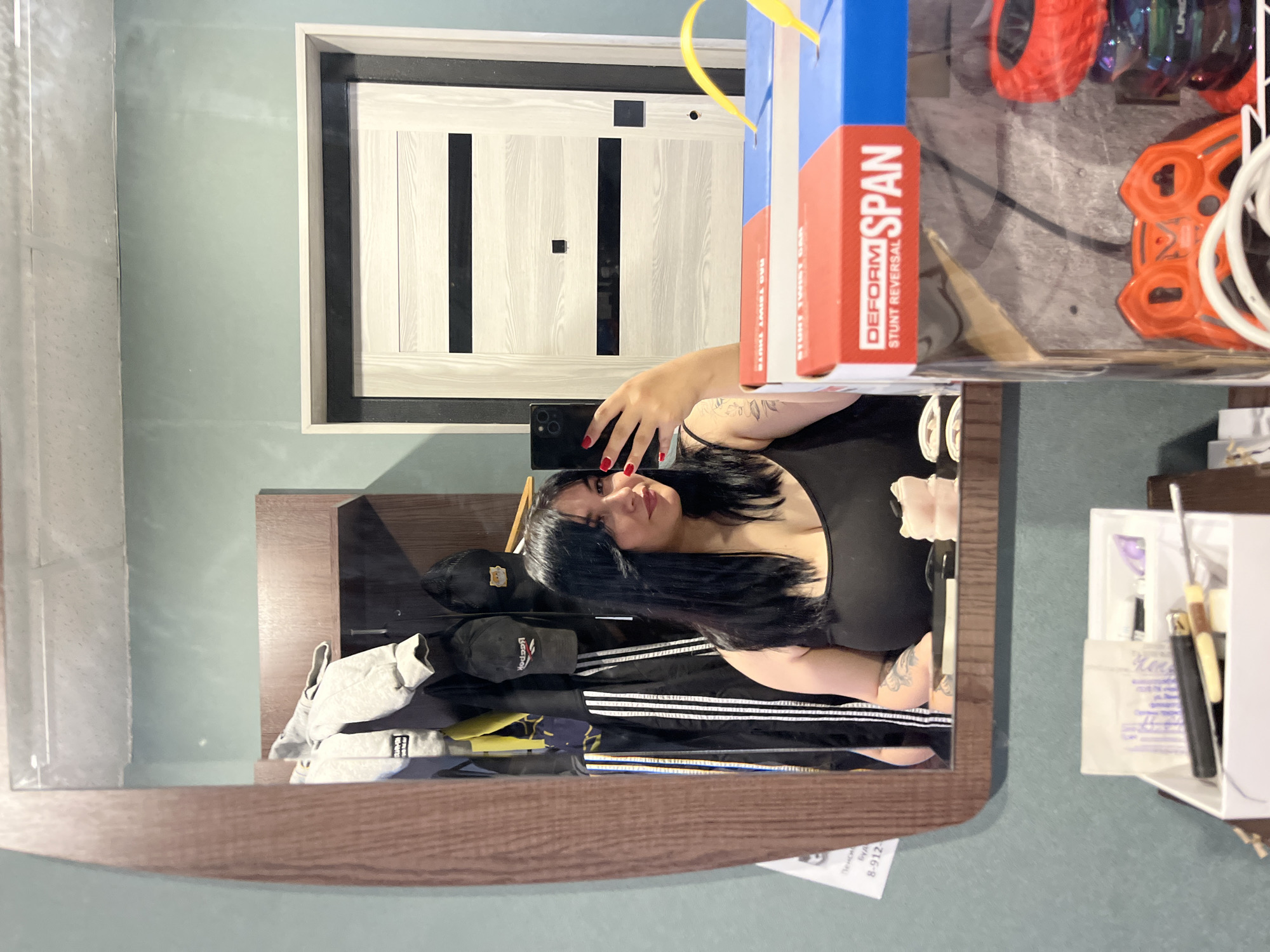 A person is taking a selfie in a mirror. The mirror is reflecting a room with a door, clothes on a shelf, and a box. The box is labeled "DEFORM SPAN STUNT TWIST CAR." The person has dark hair, red nail polish, and is wearing a black top. A piece of paper is in the lower right corner which includes phone number 8-912. No location data is confirmed in the image. The location in the city of Chaikovsky, Russia is speculative and unconfirmed.