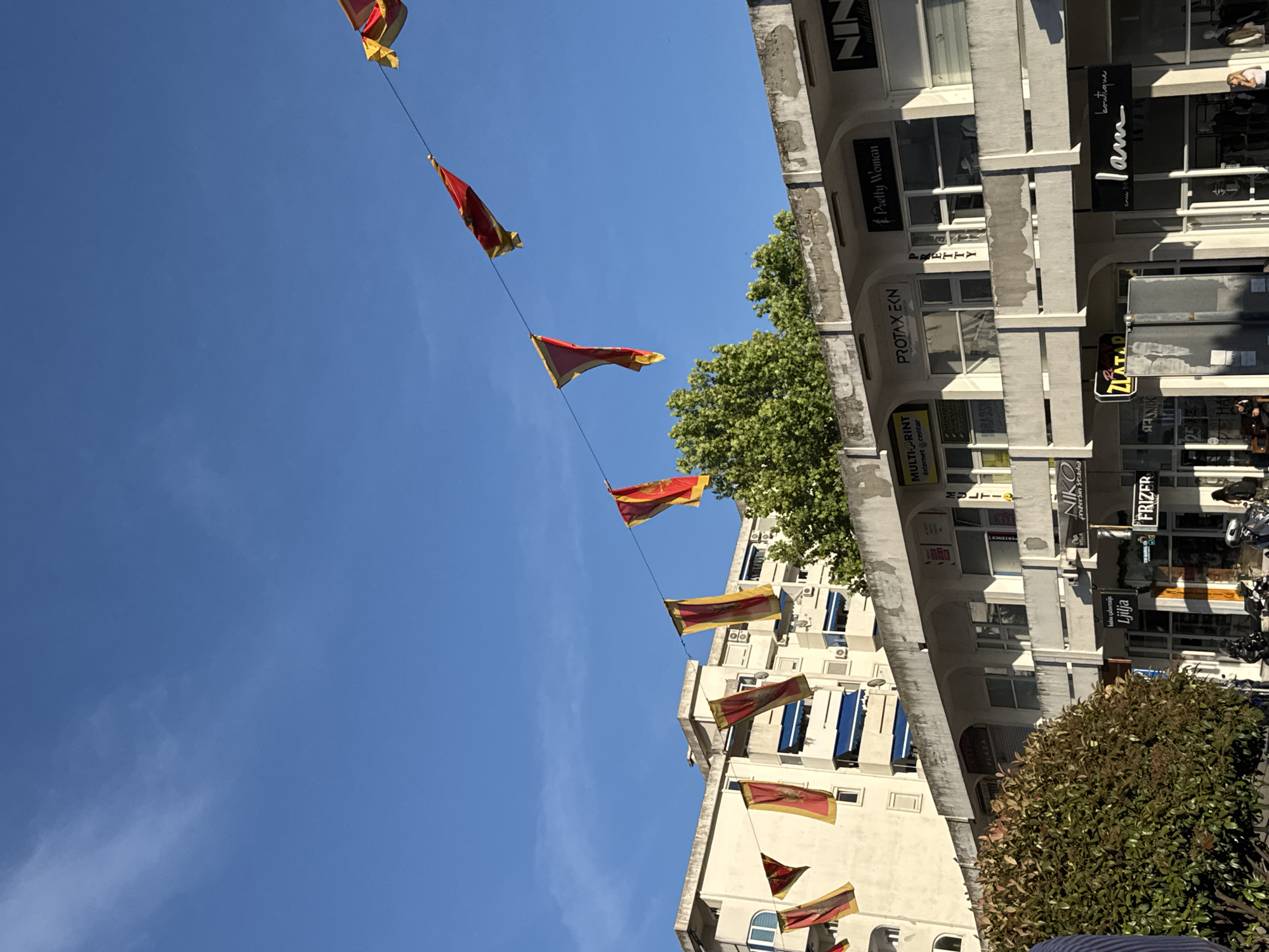 The image captures a street scene in Bar, Montenegro, with a clear blue sky overhead. A series of Montenegrin flags are strung across the street on a wire, hanging above the buildings. The buildings are predominantly white with repeating architectural patterns, including arched windows and balconies. Storefronts are visible, with signage for businesses such as "Pretty Woman," "FRIZER," and "MULTIPRINT." There are trees and shrubbery visible, some of which are obscuring the base of the buildings. A few people are visible in the storefronts or walking along the street. The overall impression is that of a bright, sunny day in an urban area.