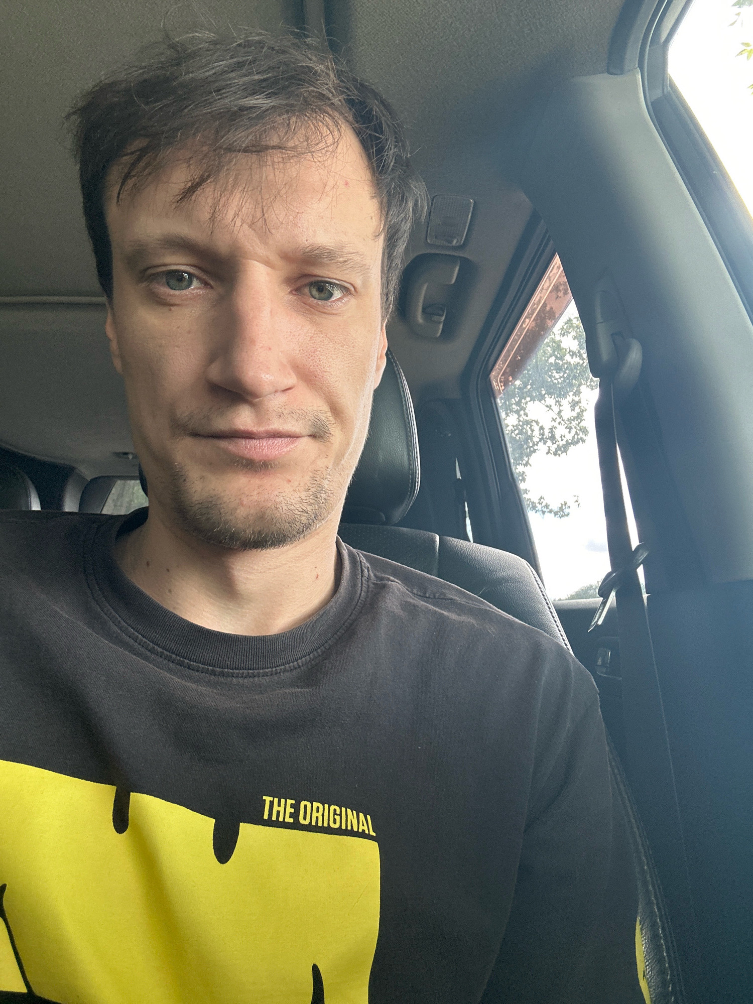A man is pictured inside a vehicle, likely a car. He is wearing a black t-shirt with a large yellow graphic design and text that reads "THE ORIGINAL". He has short, dark hair and is looking directly at the camera. The interior of the car is visible, including the seats and the window. Outside the window, trees and sky can be seen, suggesting an outdoor setting. The image appears to be taken in the city of Makiv, Ukraine.