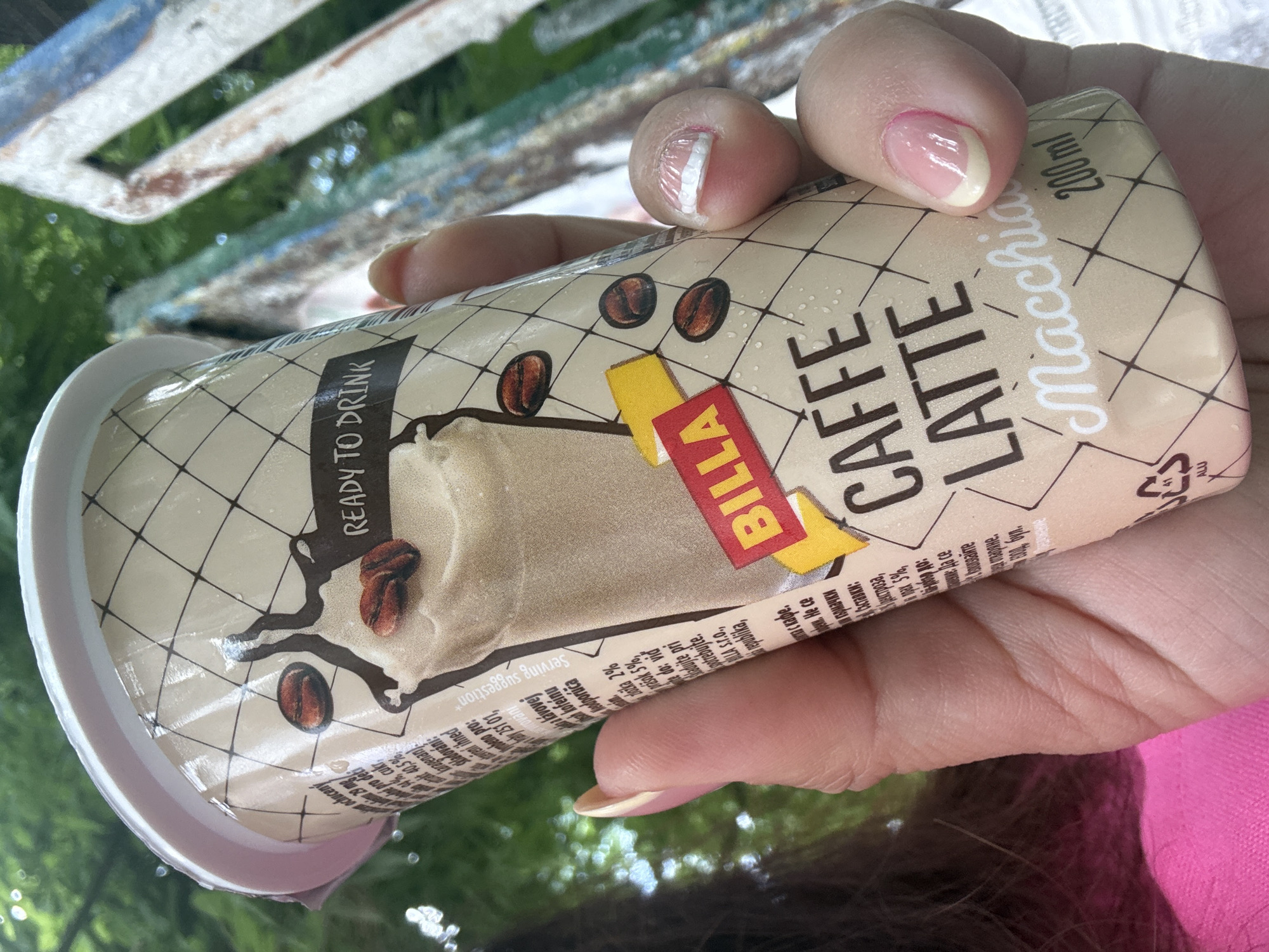 A close-up, eye-level shot shows a person's hands holding a light brown disposable cup with a white lid. The cup has a graphic design featuring coffee beans, a stylized world map, and text. Prominently displayed on the cup are the words "CAFFE LATTE" in large, dark brown capital letters, and "Macchiato" in white cursive script. Above "CAFFE LATTE," a yellow and red rectangular label reads "BILLA." Text in a smaller font is also visible on the cup, with some of it cut off by the frame. The background is blurred, with hints of green foliage and what appears to be a weathered, blue and white structure. The person's fingers, with light pink and beige nail polish, are visible on the sides of the cup. The overall impression is a casual, outdoor setting.