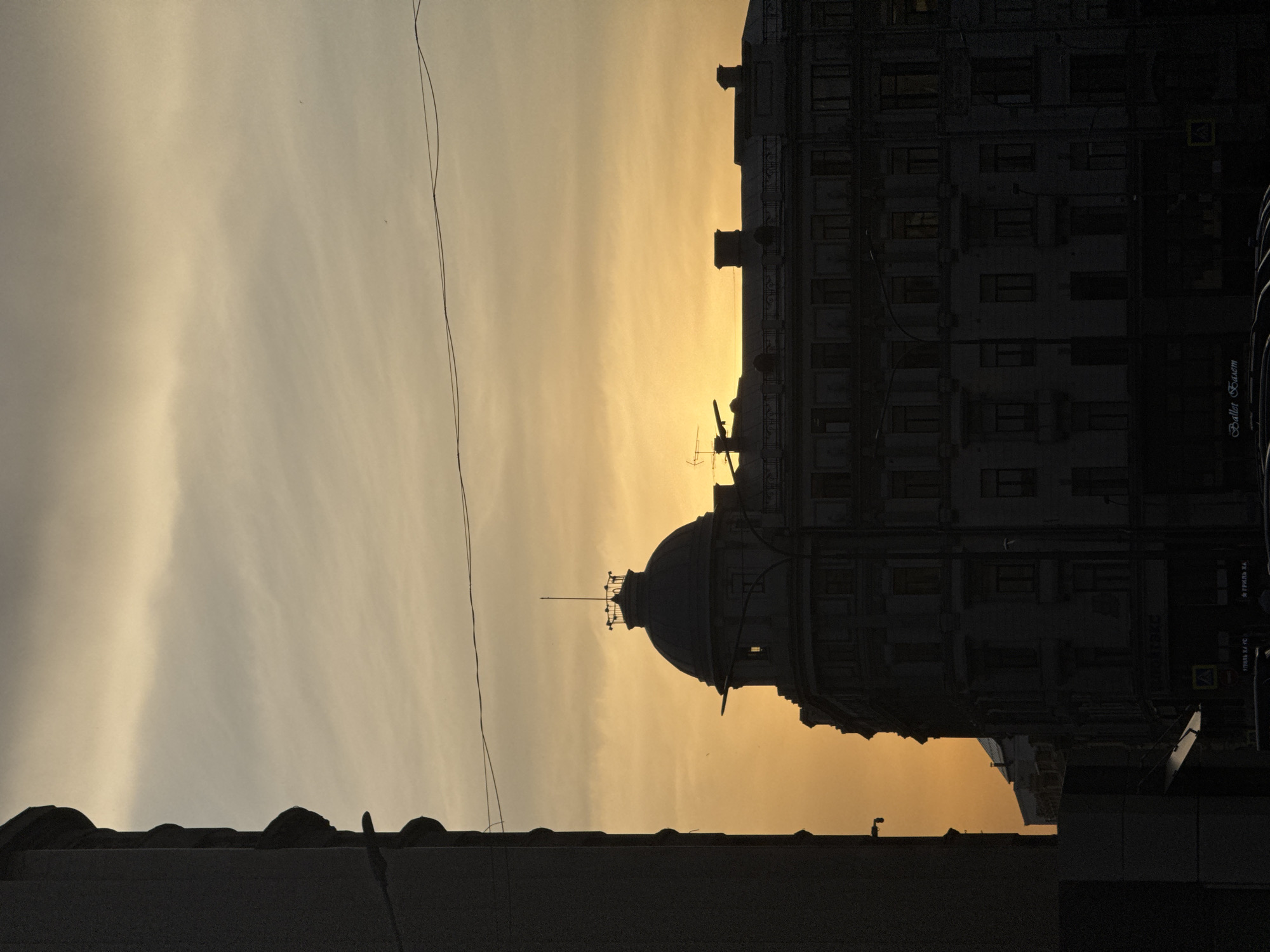 The image is a street-level view in Moscow, Russia, taken during sunset. Dominating the frame is the silhouette of a building with a prominent dome and a weather vane. To the right, another taller building is visible, with multiple windows and architectural details. A sign with Cyrillic script is also discernible on the right-hand building. In the foreground, part of a lower building is visible, creating depth. Power lines run across the top part of the sky. The sky itself has a gradient of colors, indicative of a sunset, from light gray to deep orange.