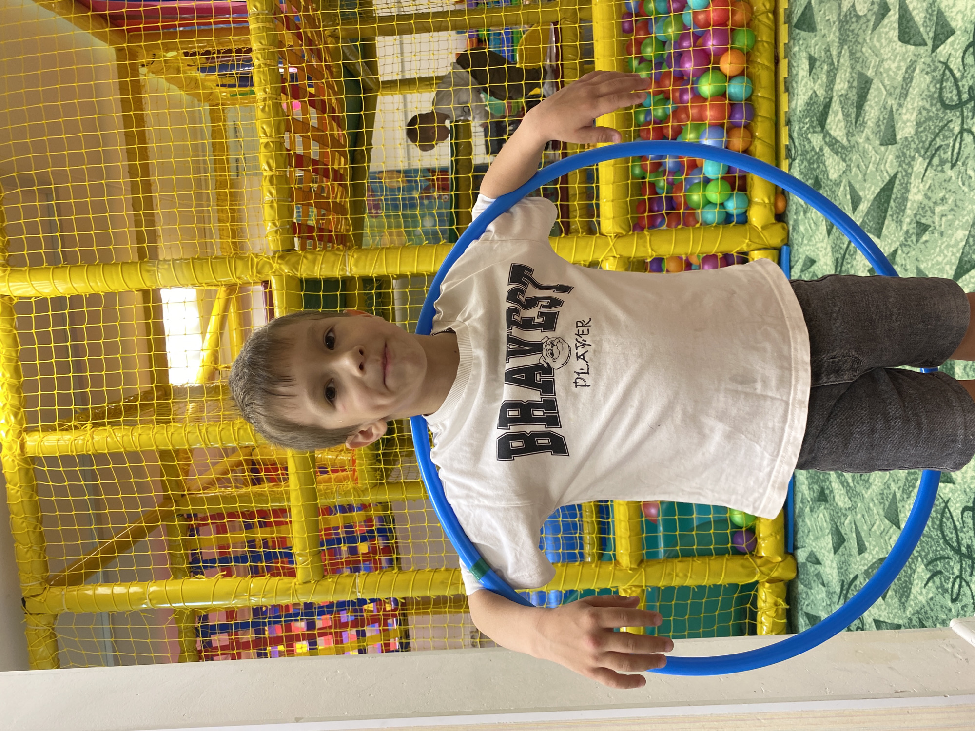 A boy stands inside a play area with a blue hula hoop around his torso. The play area has yellow netting, colorful balls, and other play equipment. He wears a white t-shirt with the word "BRAVES" printed on it and gray shorts. The floor is white with patterned wallpaper in the background. The location is Chaikovsky, Russia.