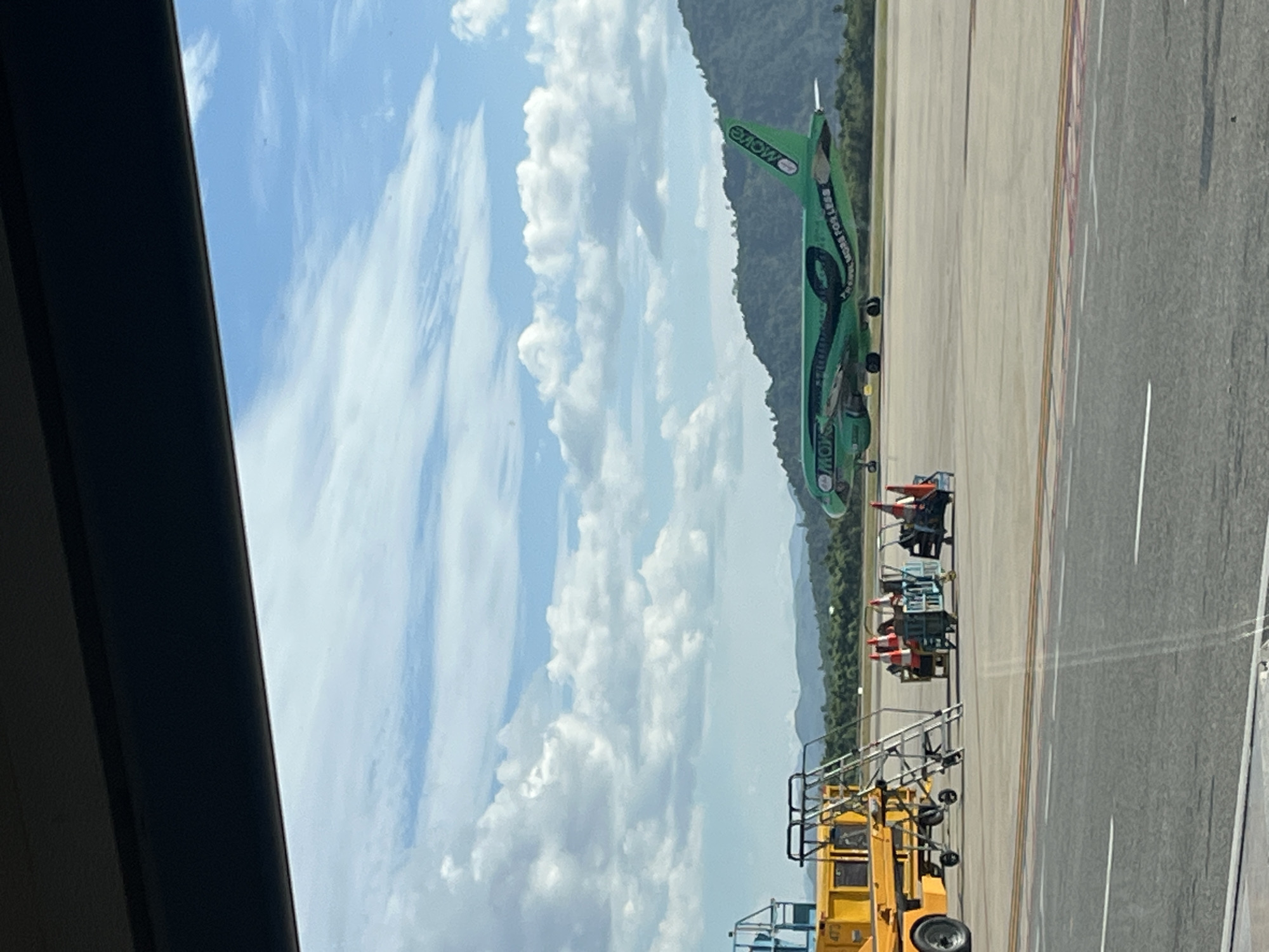 This image depicts an airport apron scene at Đà Nẵng International Airport in Đà Nẵng, Vietnam, viewed through a window with a dark frame at the top.
In the mid-ground, a bright green commercial aircraft, identified as a Vietjet Air plane by the "MOWO" logo on its tail and "MORE FOR LESS" text on its fuselage, is parked or taxiing on the tarmac. The twin-engine jet is facing generally towards the right, with its tail slightly angled towards the left.
To the left of the aircraft, several pieces of ground support equipment are visible: a yellow wheeled vehicle with blue components (partially obscuring a number, "473"), a silver metallic mobile staircase, and two blue trolleys laden with orange traffic cones.
The foreground consists of a dark gray tarmac with white and orange markings, indicating airport operational areas. The background features a long, light gray runway or taxiway, beyond which lies a range of lush green, vegetation-covered mountains under a bright blue sky filled with numerous white cumulus clouds. No people are distinctly visible in the immediate vicinity of the aircraft or ground equipment.