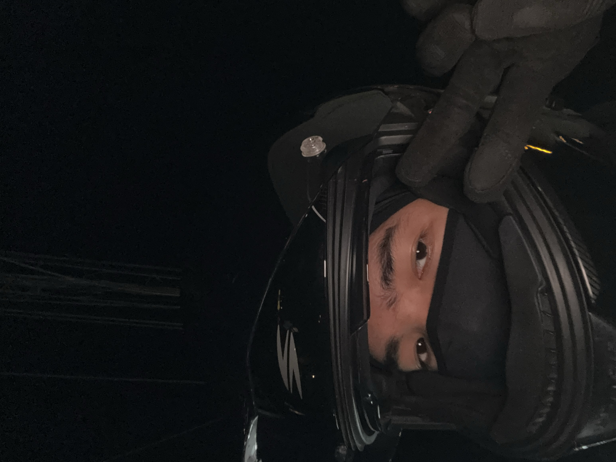 A person wearing a black motorcycle helmet and black face mask poses for a selfie in a dark environment. The helmet has a white logo on the side. The person's eyes are visible above the mask. Their right hand, wearing a black glove, is raised and covers part of the helmet. In the background, faint vertical lines suggest structures or a fence. The image appears to have been taken at night. There is no specific indicator of the location being in Davao City, Philippines.