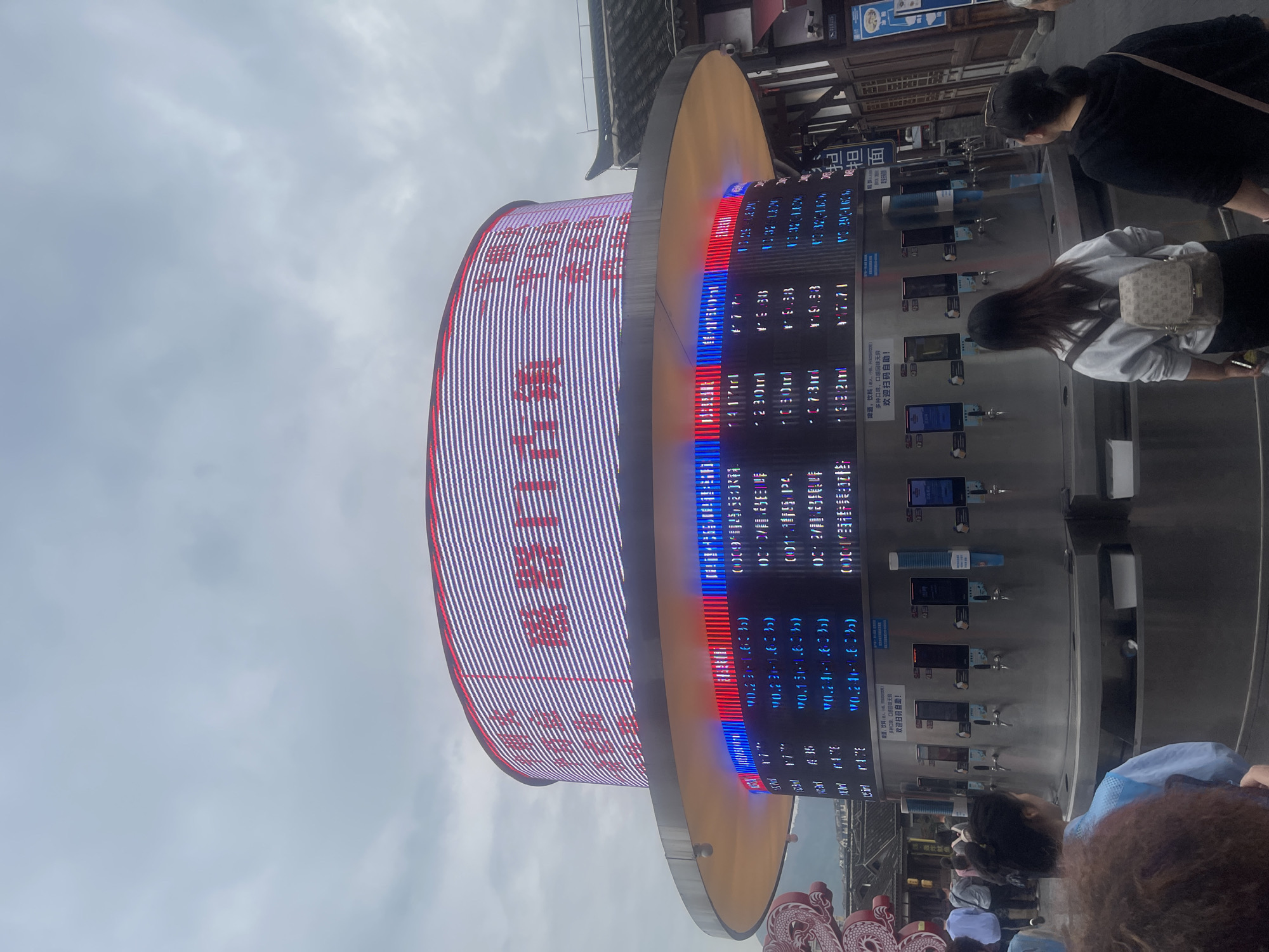 The image shows a public space in Shapingba District, China. The sky is overcast. A large circular sign dominates the frame, displaying what appears to be stock market information in Chinese characters and numbers. The sign has red and blue accents. Below the sign, people are gathered. Some are interacting with vending machines that dispense beverages. The vending machines have digital displays and dispensing spouts. The background includes architectural elements with traditional Chinese design.