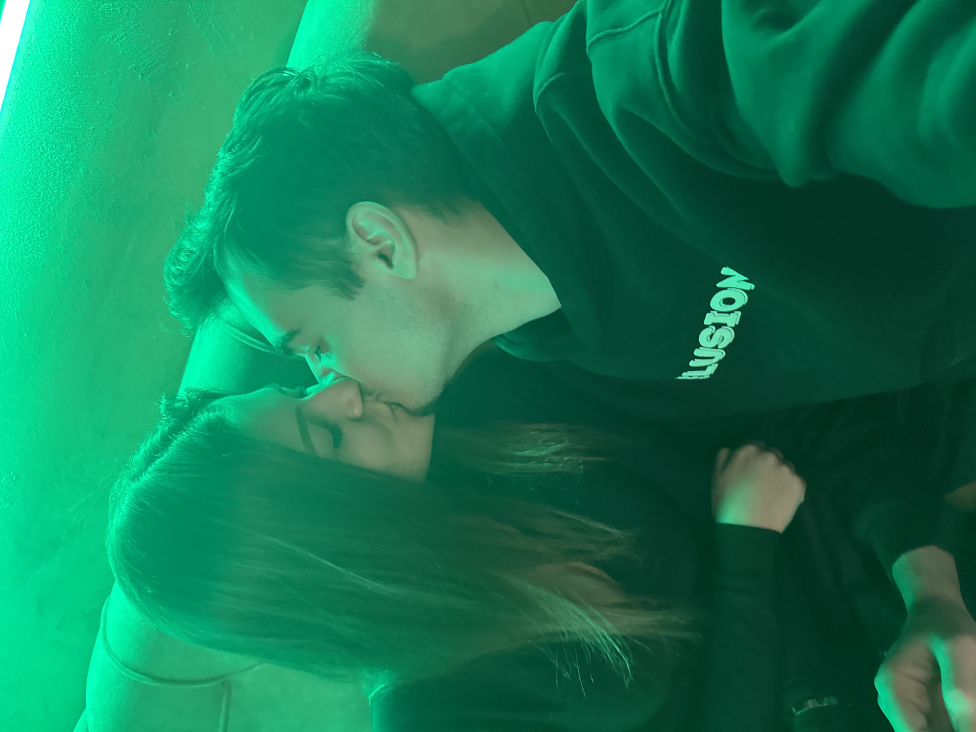 A photograph depicts a man and a woman kissing indoors under a green light. The man wears a black hooded sweatshirt with the word "LUSION" visible on the front. The woman has long brown hair. The location is unknown and there is no evidence of the image being taken in Sofia, Bulgaria.