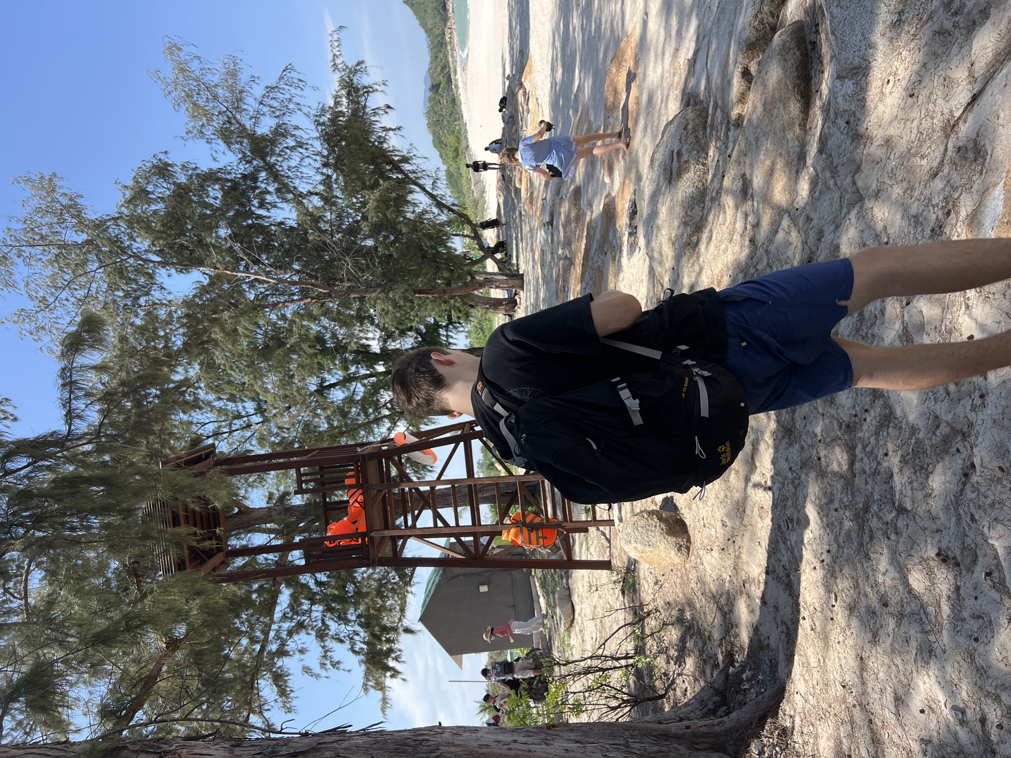This image depicts a coastal scene in Cong Hai, Vietnam, under a bright, clear blue sky. The foreground is dominated by a male figure, viewed from behind, wearing a black t-shirt, dark blue shorts, and a black Jack Wolfskin backpack. He stands on a sandy, sun-dappled ground interspersed with light brown rocks and dry foliage.
To the left, a large tree trunk occupies the extreme left of the frame. Further back, a dark grey, single-story building with a light roof is partially visible, with a group of people nearby, including one person in a light pink hat. To the right of the building stands a tall, multi-tiered wooden structure, resembling a lifeguard stand or observation tower, with a ladder and a small roof. Two bright orange life vests are secured on the structure, one on the top platform and another on a lower section. A white and orange flotation device is also attached to the structure.
Beyond the foreground and midground, a sandy beach extends towards a turquoise sea on the right. A young person in a light blue dress is walking barefoot on a rocky, sandy path towards the water, carrying a pair of shoes. Further along the beach, several indistinct figures are visible: one person stands near the waterline, two are seated on rocks or the sand, and another is lying down. Green, tree-covered hills rise in the distant background, bordering the sea. The scene is illuminated by strong sunlight, creating distinct shadows on the sandy ground.